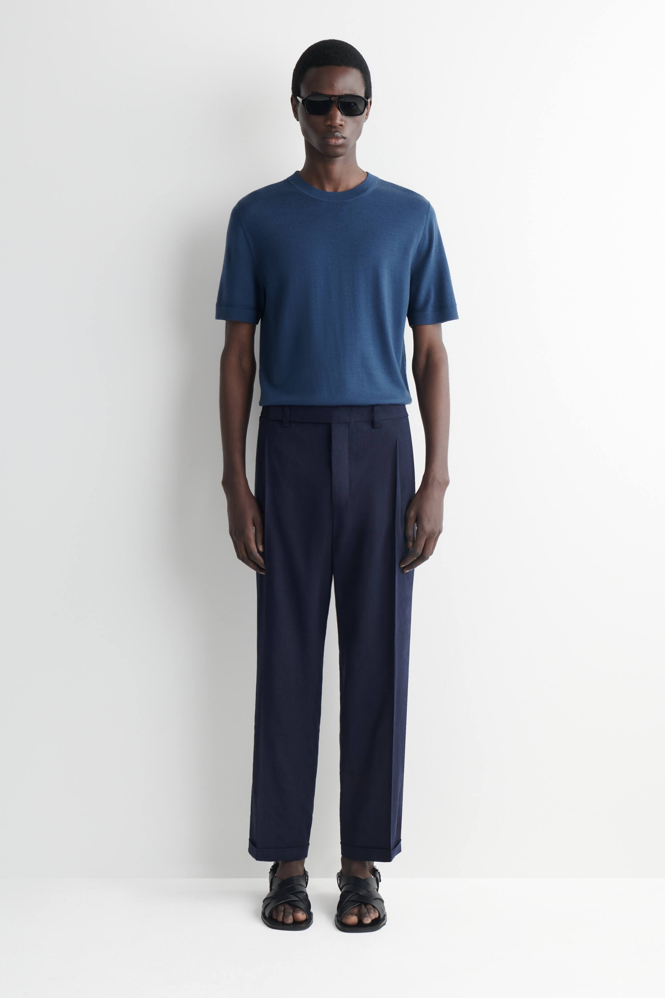 Visualizza immagine più grande: Una maglietta blu a maniche corte in maglia è indossata, infilata in pantaloni a vita alta blu navy con pieghe. I pantaloni presentano una gamba dritta e orli risvoltati, e ai piedi sono indossati sandali neri con cinturini.