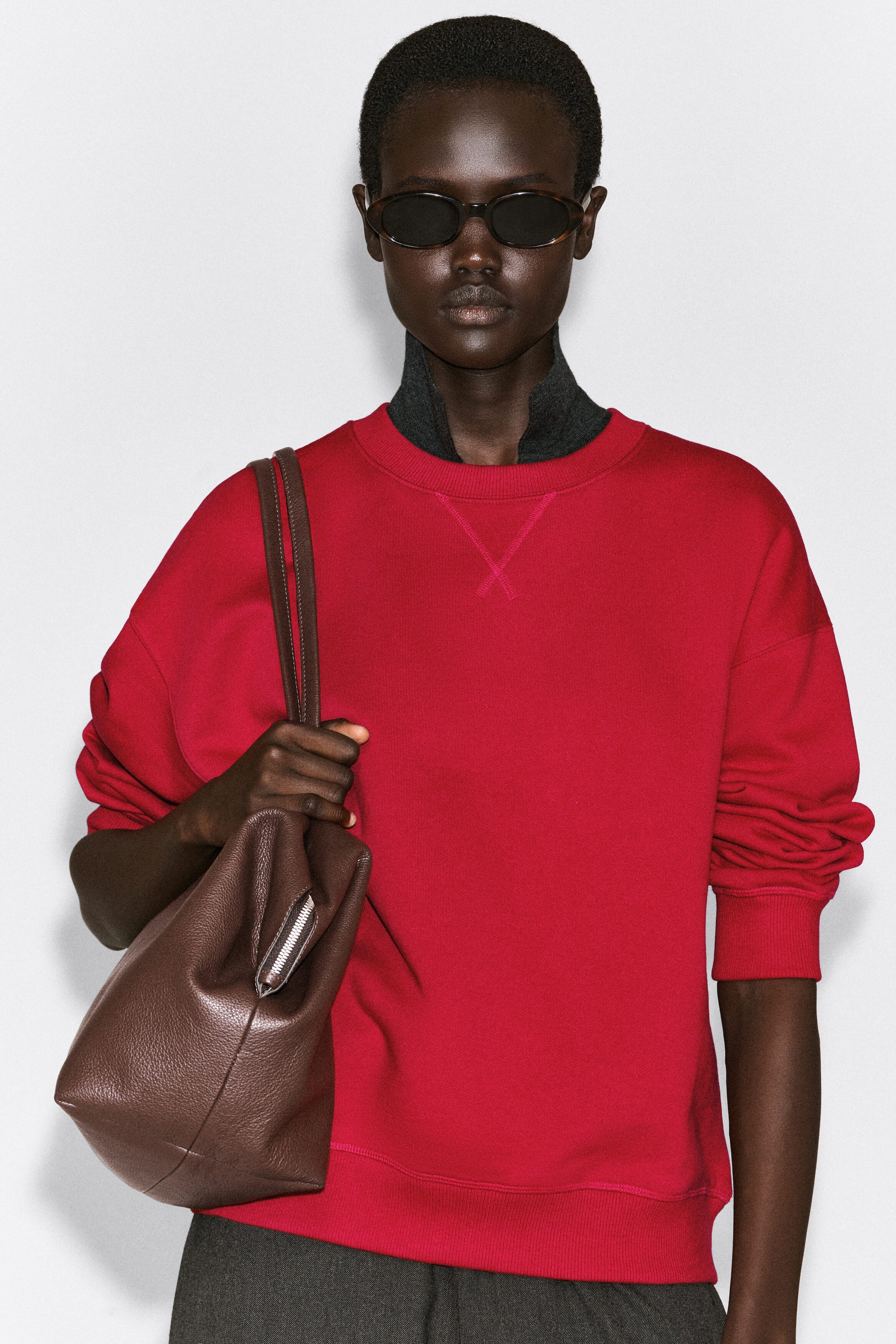 A red, rounded cotton crew-neck sweatshirt is shown in a front-facing view, featuring a relaxed fit with dropped shoulders, ribbed cuffs, and a ribbed hem. The sweatshirt features a tonal V-stitch detail below the ribbed collar and is layered over a dark gray high-neck top.