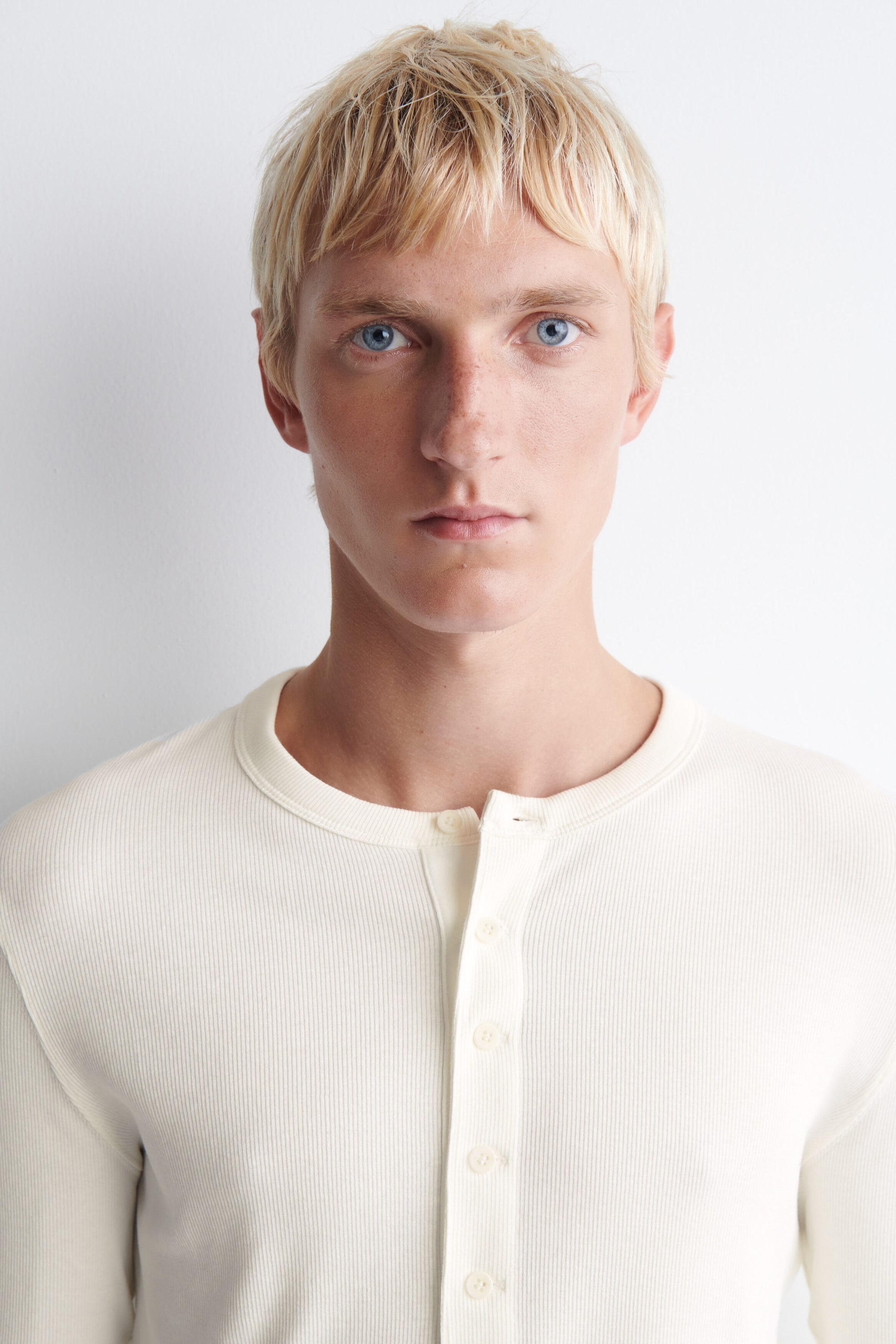 A slim-fit, off-white ribbed cotton long-sleeved Henley t-shirt is shown facing forward, highlighting its fine vertical ribbed texture and classic crew neckline. The garment features a vertical button placket down the centre chest, with the top buttons left unfastened to showcase its casual structure. A slim-fit, off-white ribbed cotton long-sleeved Henley t-shirt is shown facing forward, highlighting its fine vertical ribbed texture and classic crew neckline. The garment features a vertical button placket down the centre chest, with the top buttons left unfastened to showcase its casual structure.