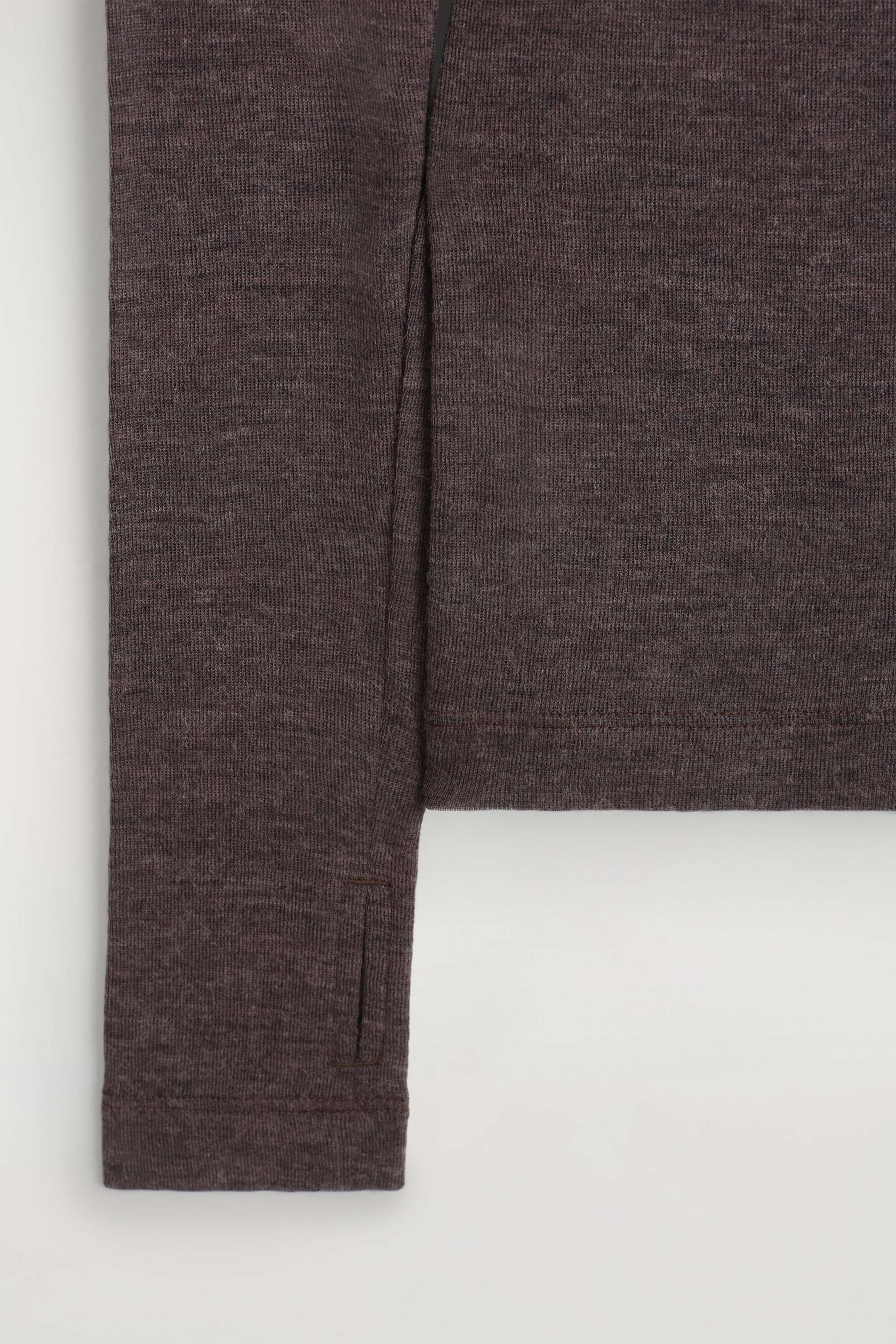 The Slim Merino Wool Top is shown in a close-up view, displaying its dark brown melange, fine-knit texture. The long sleeve extends downward, featuring a stitched thumbhole detail near the cuff beside the straight hem of the body. The Slim Merino Wool Top is shown in a close-up view, displaying its dark brown melange, fine-knit texture. The long sleeve extends downward, featuring a stitched thumbhole detail near the cuff beside the straight hem of the body.