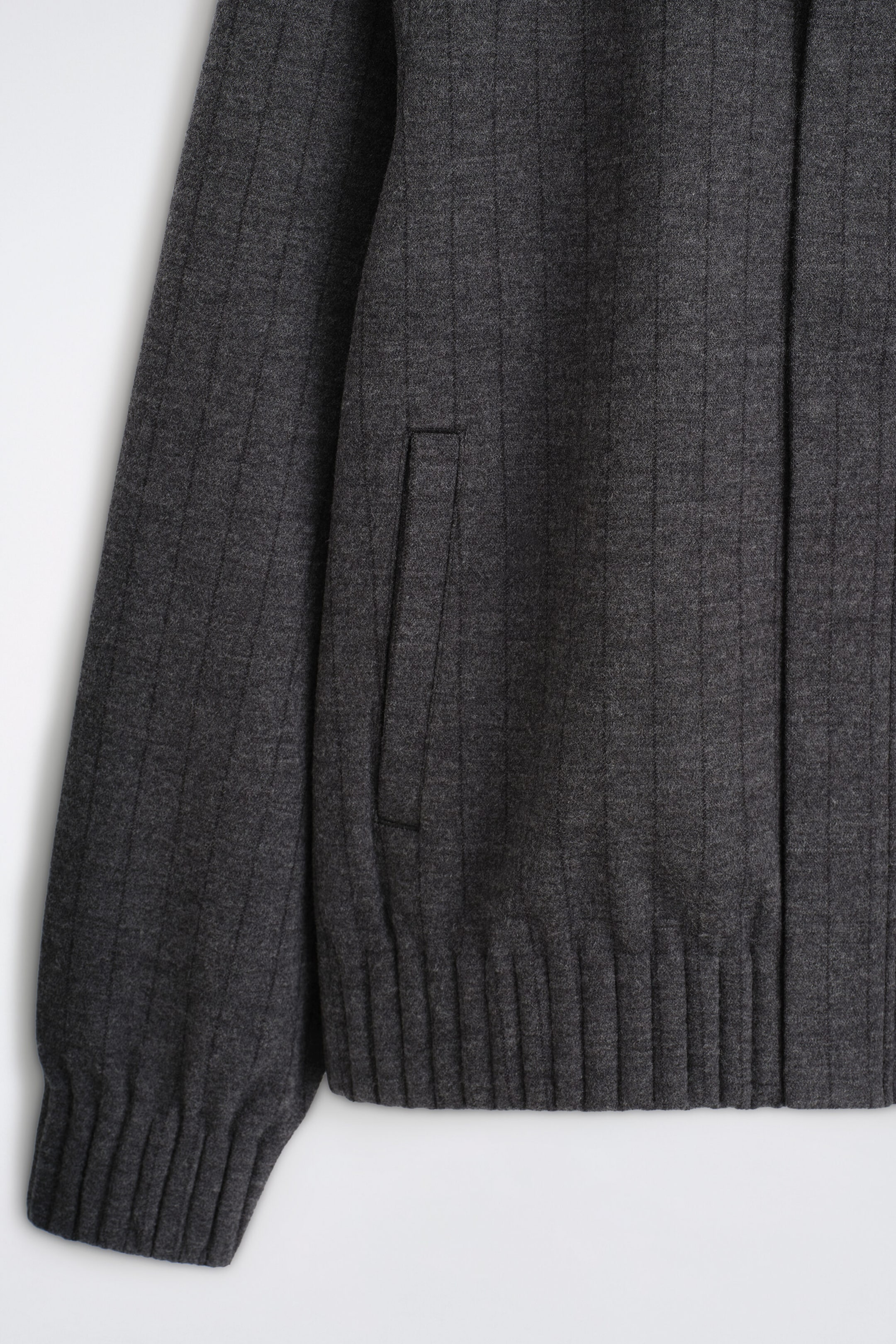 A dark gray jacket is shown in a close-up front view, featuring a vertical ribbed texture, a vertical welt pocket, a ribbed hem, and a ribbed cuff on the sleeve. A dark gray jacket is shown in a close-up front view, featuring a vertical ribbed texture, a vertical welt pocket, a ribbed hem, and a ribbed cuff on the sleeve.