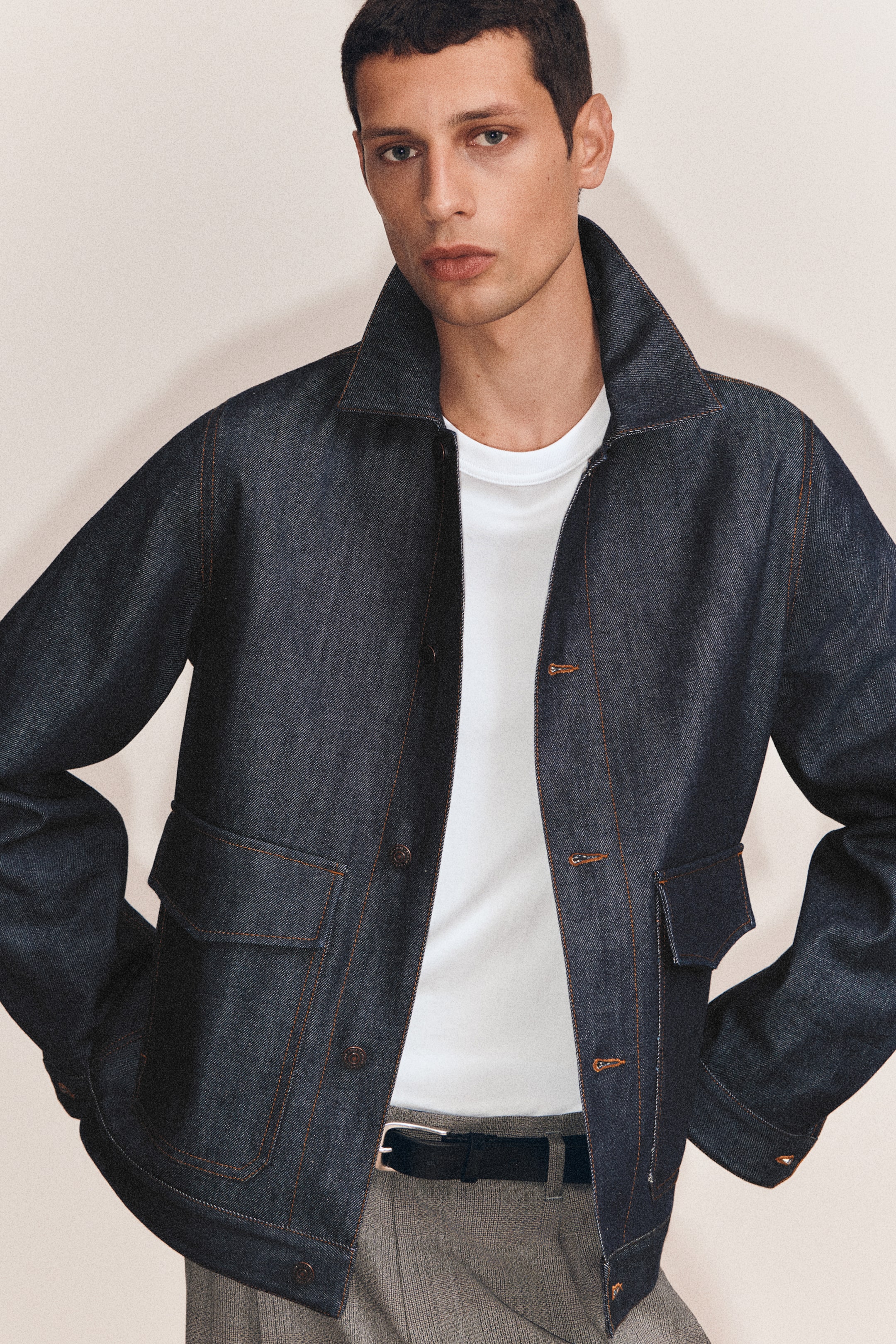 Agrandir l'image: Une veste-chemise en denim bleu foncé est présentée de face, mettant en valeur une coupe décontractée, une fermeture à boutons foncés et des surpiqûres couleur tabac sur l’ensemble du modèle. Elle comporte deux poches plaquées à rabat à l’avant, en bas, et est portée sur un t-shirt blanc.