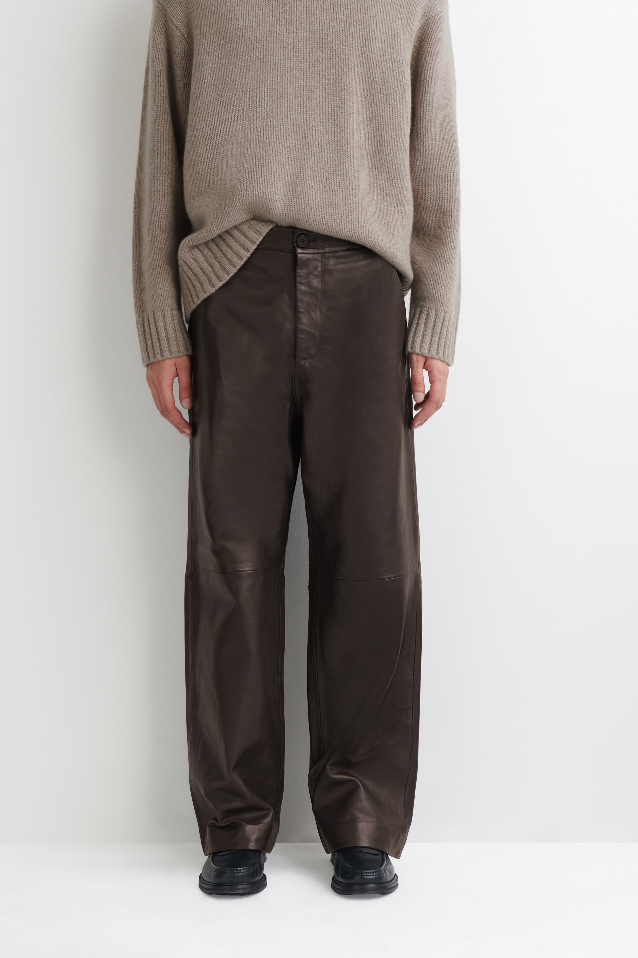 These dark brown leather relaxed-fit straight-leg pants feature a loose, straight-leg silhouette with horizontal seam detailing across the knees. The pants have a button-and-fly closure at the waistband, which is partially revealed by a tucked-in beige knit sweater. These dark brown leather relaxed-fit straight-leg pants feature a loose, straight-leg silhouette with horizontal seam detailing across the knees. The pants have a button-and-fly closure at the waistband, which is partially revealed by a tucked-in beige knit sweater.