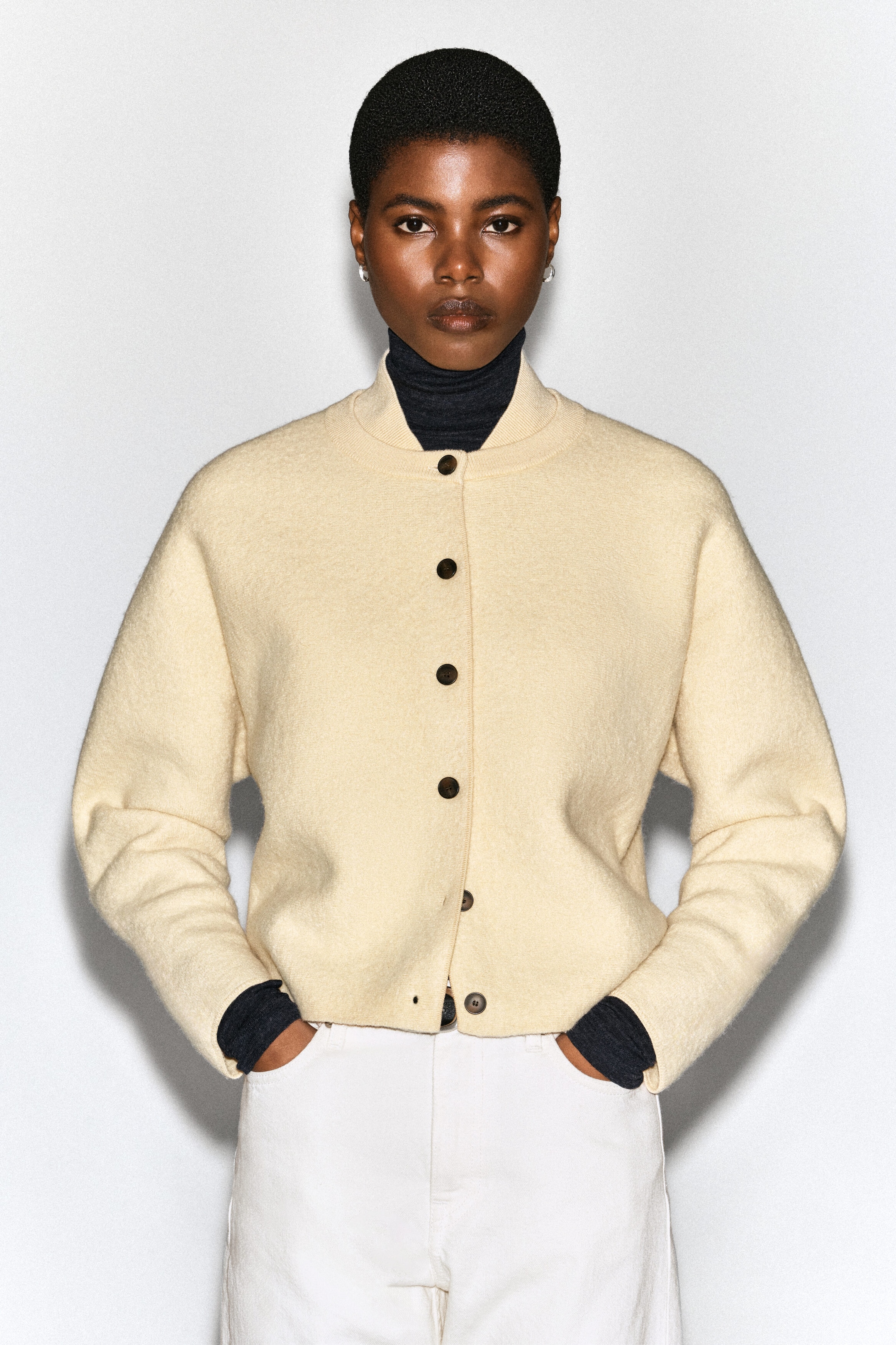 Un cardigan en laine bouillie de couleur crème, doté d’un col de style bomber et de boutons foncés, est porté par le mannequin. Le cardigan est superposé à un col roulé gris foncé et agencé avec un pantalon blanc, tandis que le mannequin se tient de face, les mains dans les poches. Un cardigan en laine bouillie de couleur crème, doté d’un col de style bomber et de boutons foncés, est porté par le mannequin. Le cardigan est superposé à un col roulé gris foncé et agencé avec un pantalon blanc, tandis que le mannequin se tient de face, les mains dans les poches.