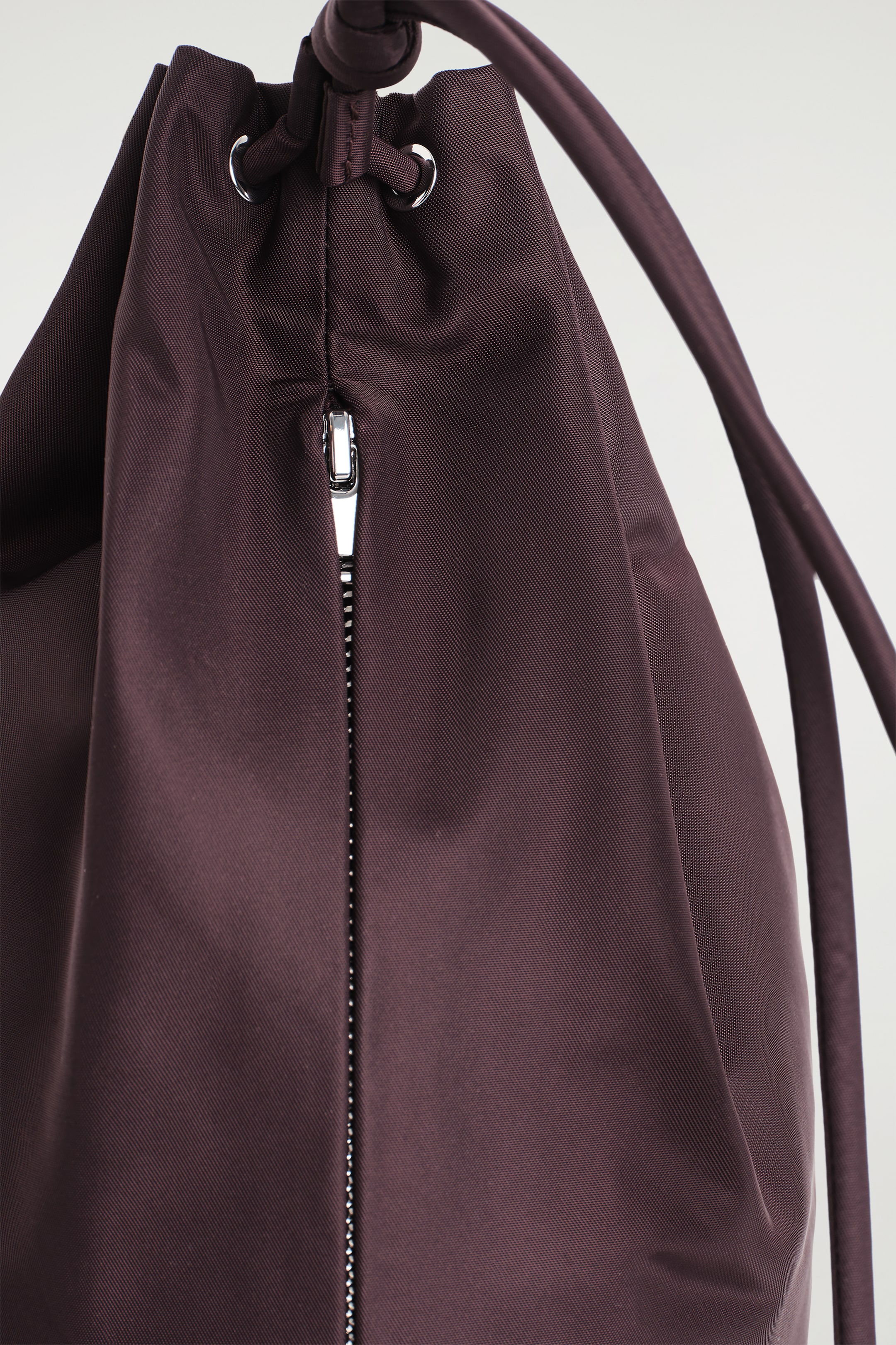 View larger image: A dark plum woven fabric drawstring bag is featuring a cinched top with silver-toned eyelets and a matching fabric cord. A partially unzipped, silver-toned vertical zipper is running down the side of the bag.