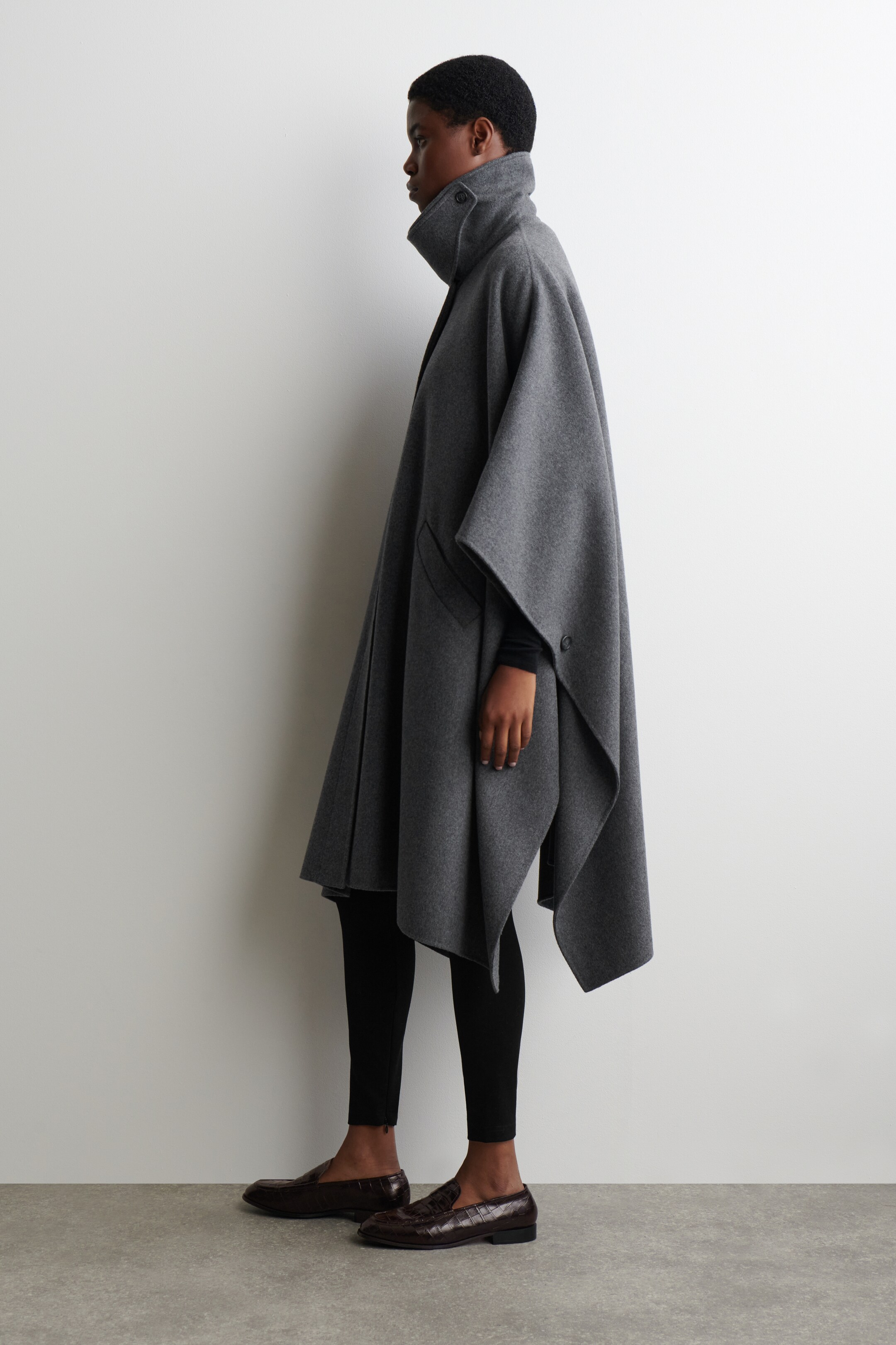 Agrandir l'image: Une cape grise est présentée de profil, dotée d'un col droit haut avec une seule fermeture à bouton. Elle présente une coupe ample et fluide avec un ourlet asymétrique et une poche plaquée visible, drapée sur des manches longues noires et des leggings.