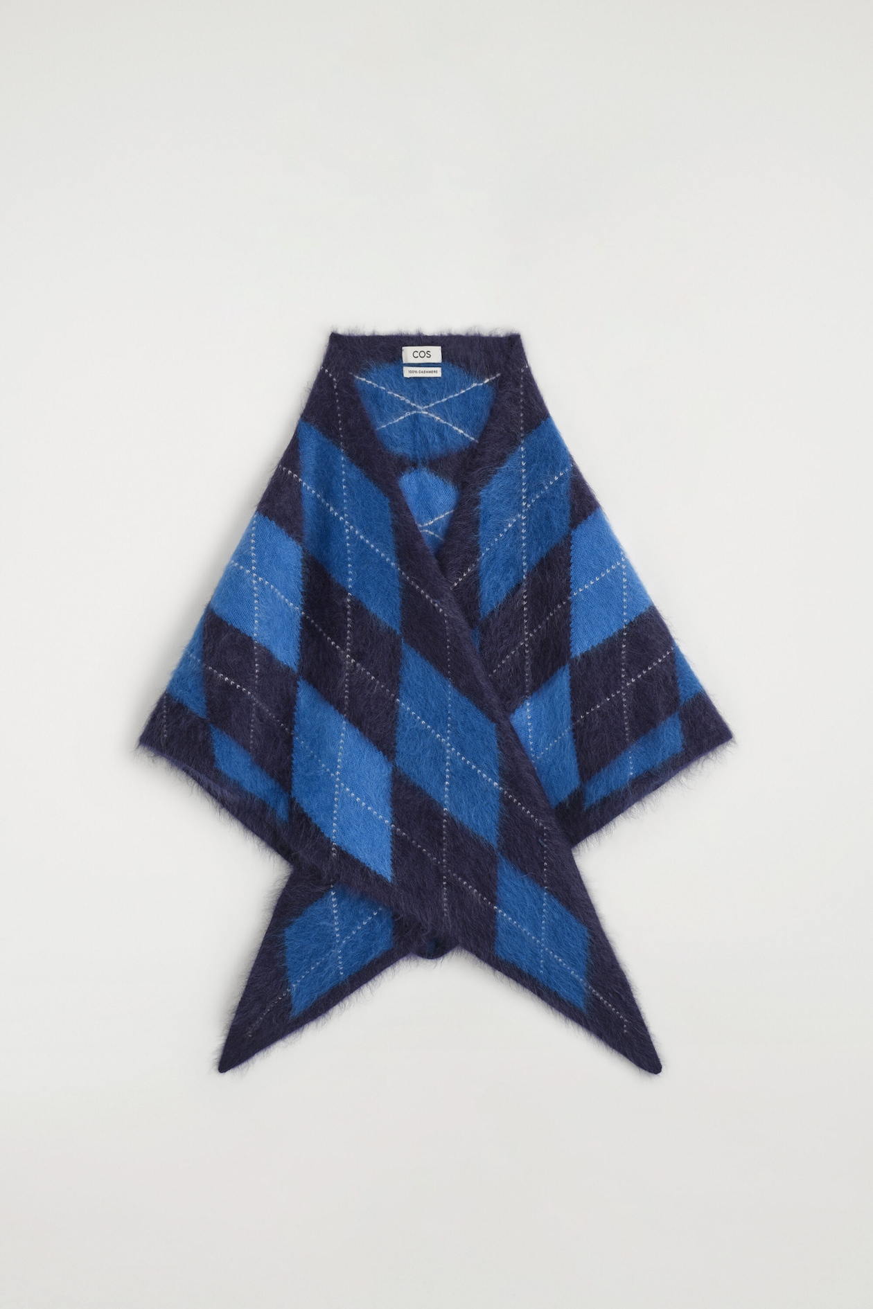 An argyle brushed cashmere triangle scarf is shown from the front, displaying a fluffy, brushed texture with a bold argyle pattern in navy and bright blue. The scarf is draped flat to highlight its triangular shape, with the pointed ends crossing over at the bottom.
