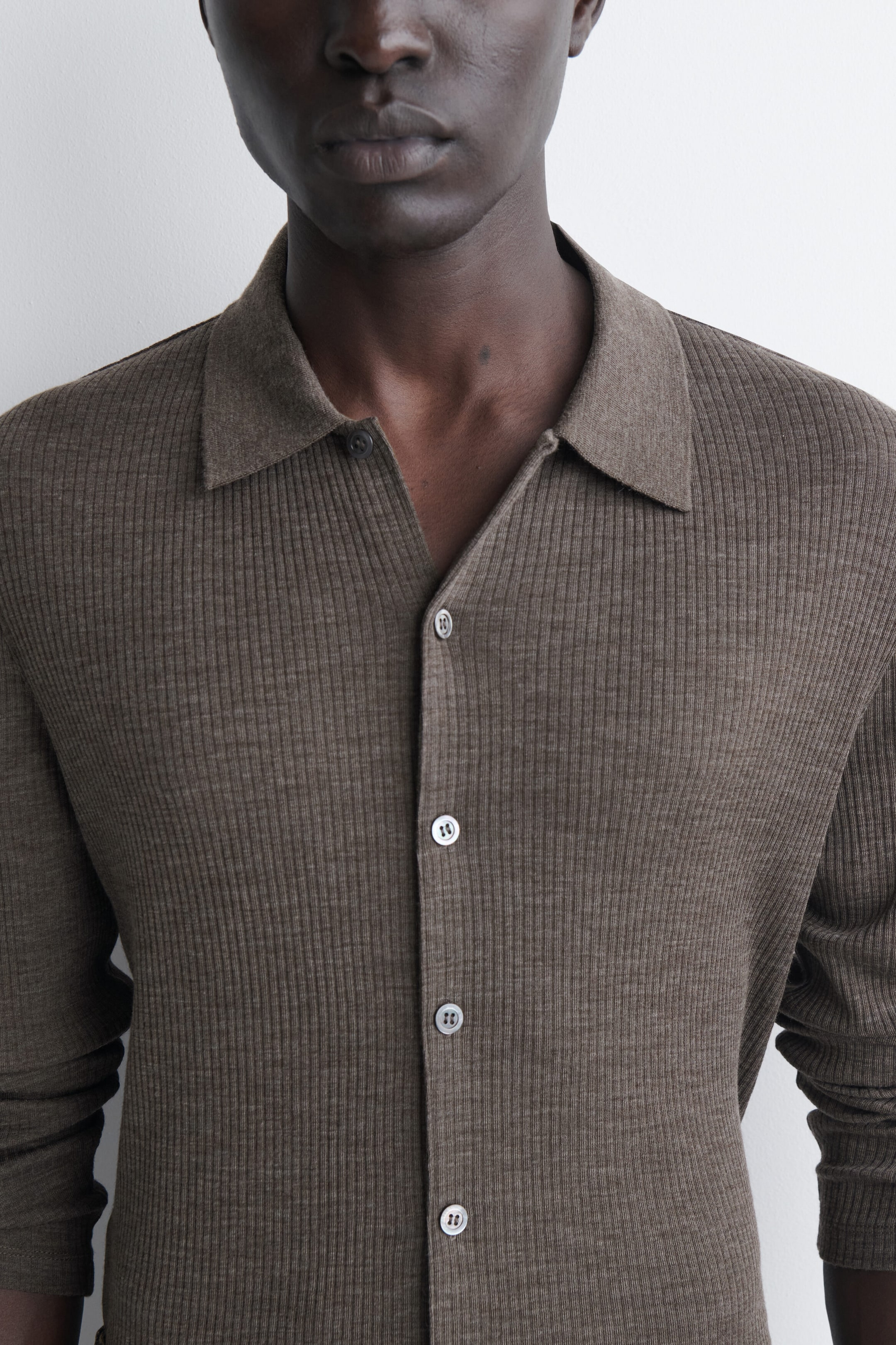 Das schmal geschnittene Wollshirt in Braun wird in einer nahen, frontalen Ansicht gezeigt, um seine figurbetonte Silhouette und die feine vertikale Rippstruktur hervorzuheben. Es verfügt über einen klassischen Flachkragen, eine durchgehende Knopfleiste mit hellen Knöpfen sowie lange Ärmel. Das schmal geschnittene Wollshirt in Braun wird in einer nahen, frontalen Ansicht gezeigt, um seine figurbetonte Silhouette und die feine vertikale Rippstruktur hervorzuheben. Es verfügt über einen klassischen Flachkragen, eine durchgehende Knopfleiste mit hellen Knöpfen sowie lange Ärmel.