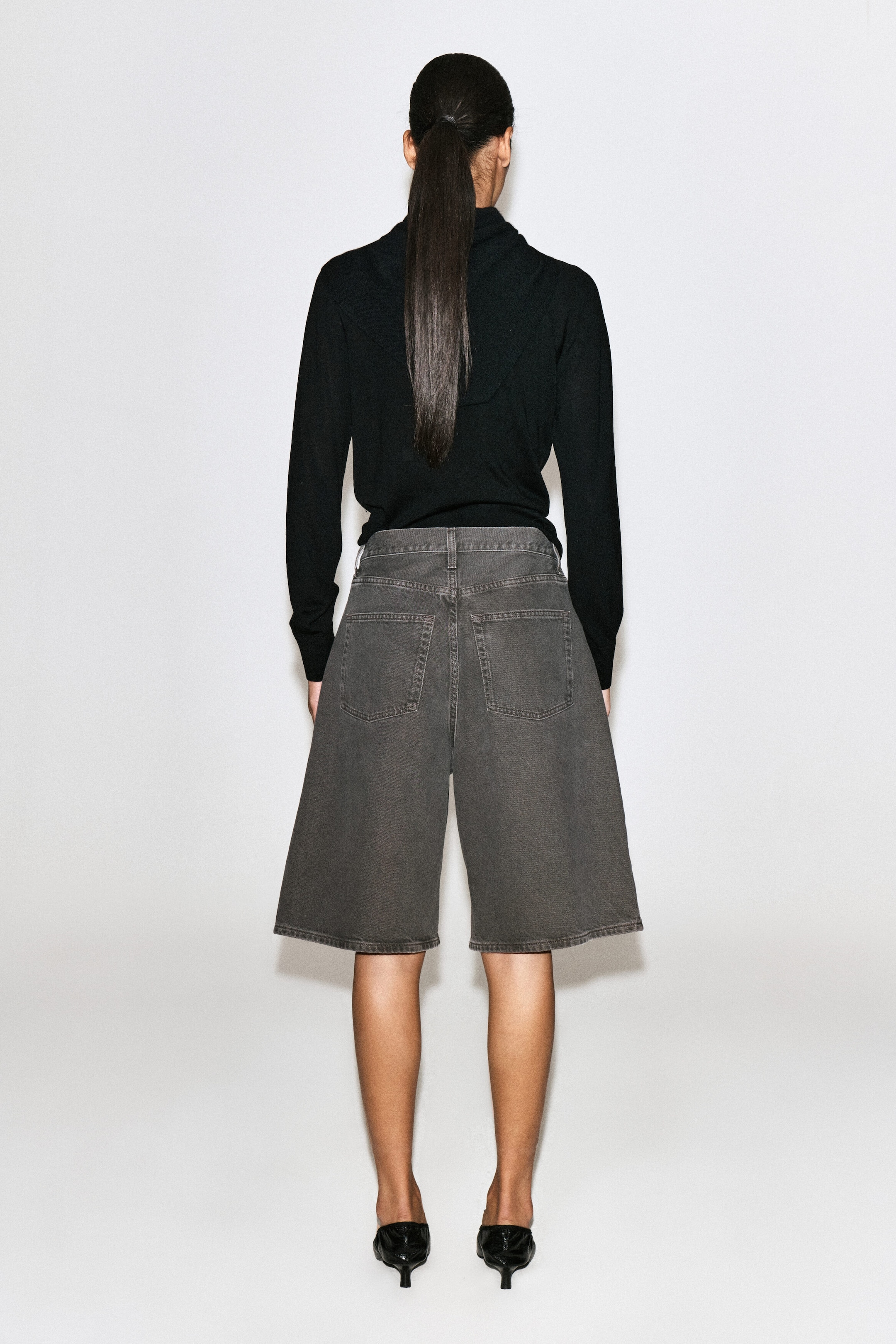 These longline denim shorts in a washed espresso hue are shown from the back, highlighting a high-rise waist, two rear patch pockets, and tonal topstitching. The wide-leg shorts fit loosely and end just above the knee, styled with a tucked-in black long-sleeved top. These longline denim shorts in a washed espresso hue are shown from the back, highlighting a high-rise waist, two rear patch pockets, and tonal topstitching. The wide-leg shorts fit loosely and end just above the knee, styled with a tucked-in black long-sleeved top.