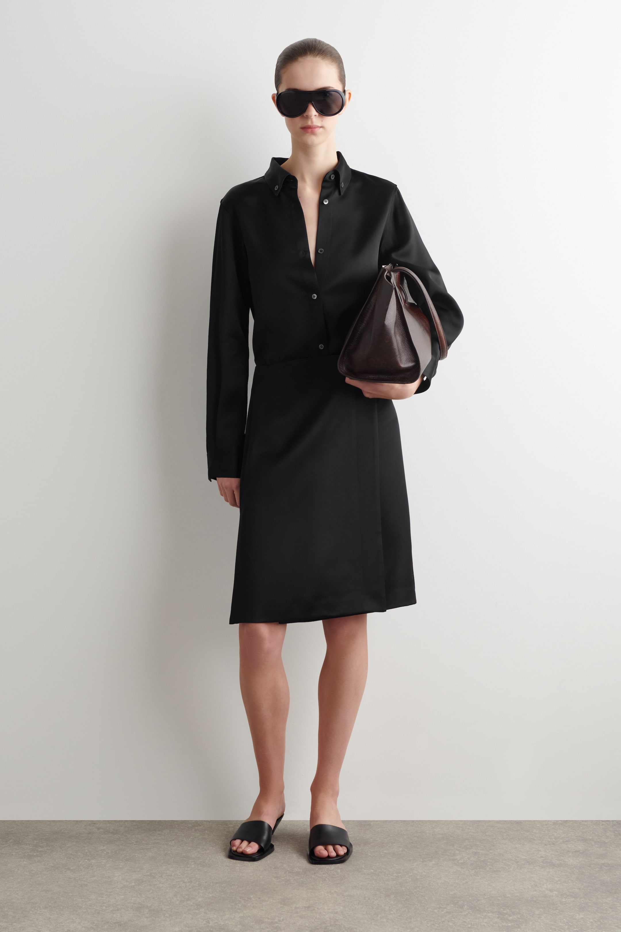 Agrandir l'image: Une jupe portefeuille en satin noir à finition douce est portée par le mannequin. Elle présente une coupe longueur genou et une fermeture asymétrique à l'avant, et est associée à une chemise en satin noir assortie.