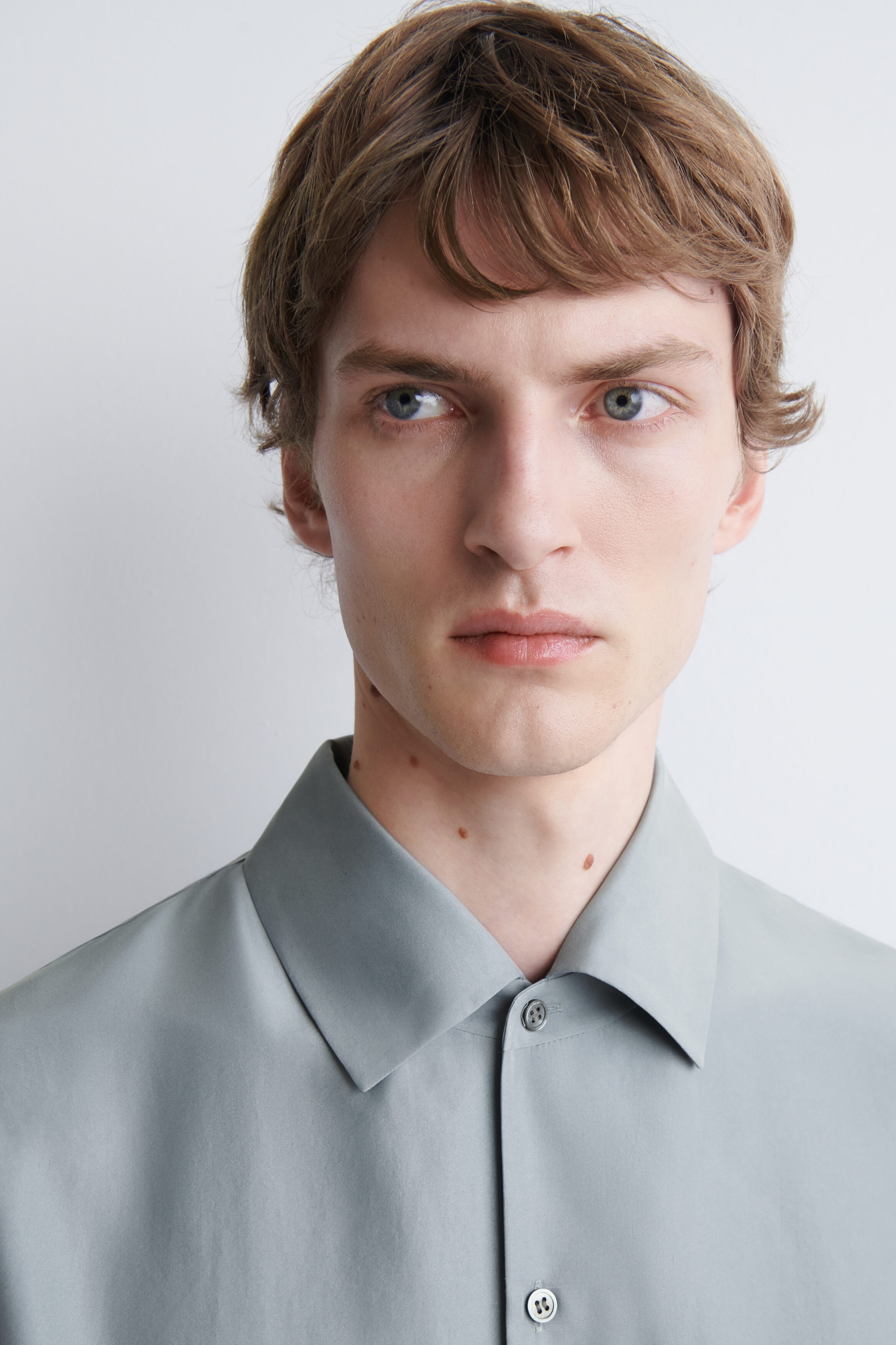 The model is wearing a relaxed silk shirt in a light sage-grey colour, featuring a classic pointed collar and a button-down front. The model is wearing a relaxed silk shirt in a light sage-grey colour, featuring a classic pointed collar and a button-down front.