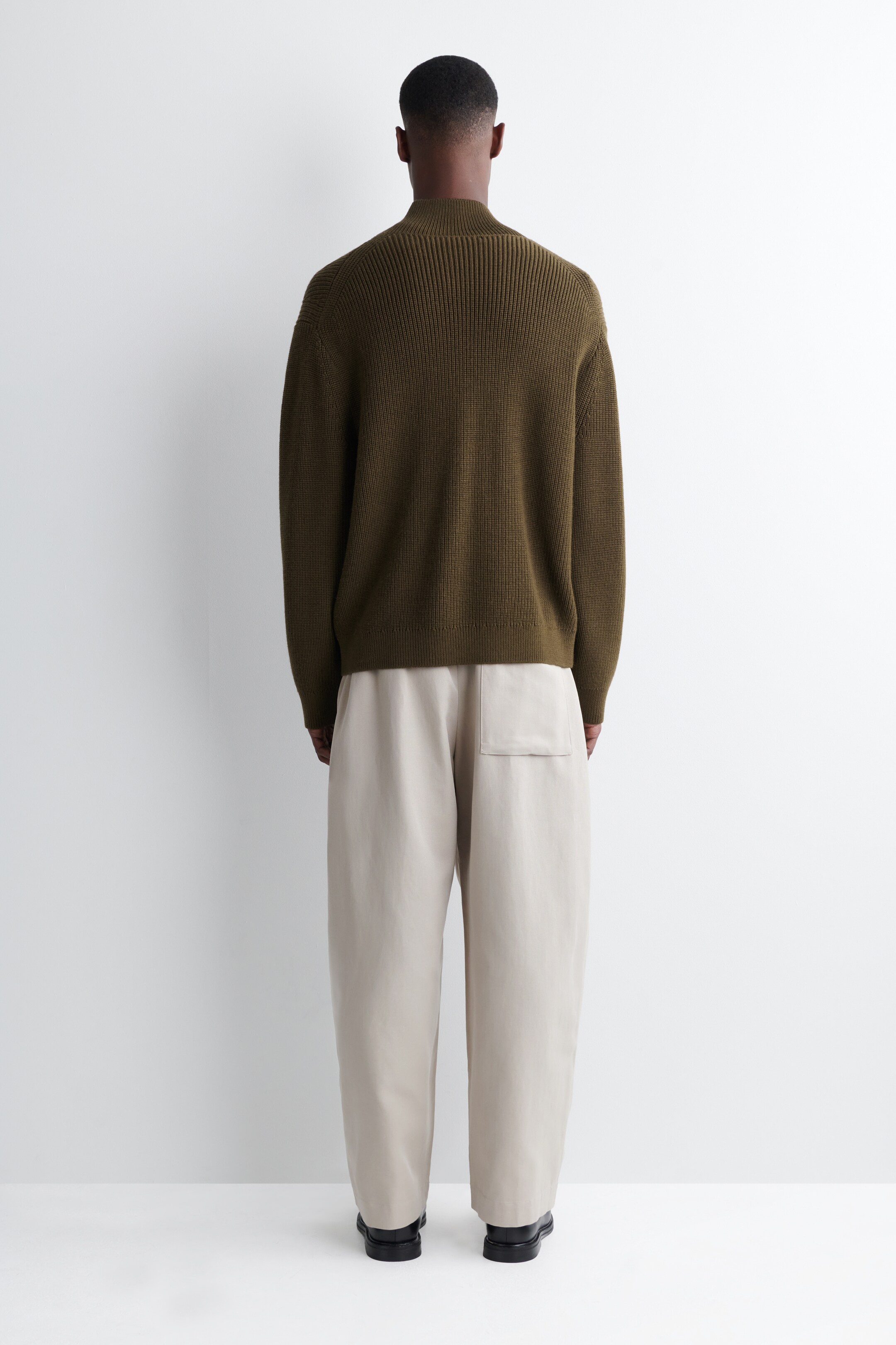 Agrandir l'image: Un pull vert olive, avec un col montant côtelé et un dos supérieur côtelé, est porté de dos. Il est assorti à un pantalon large beige clair, avec une seule poche plaquée à l'arrière droit.