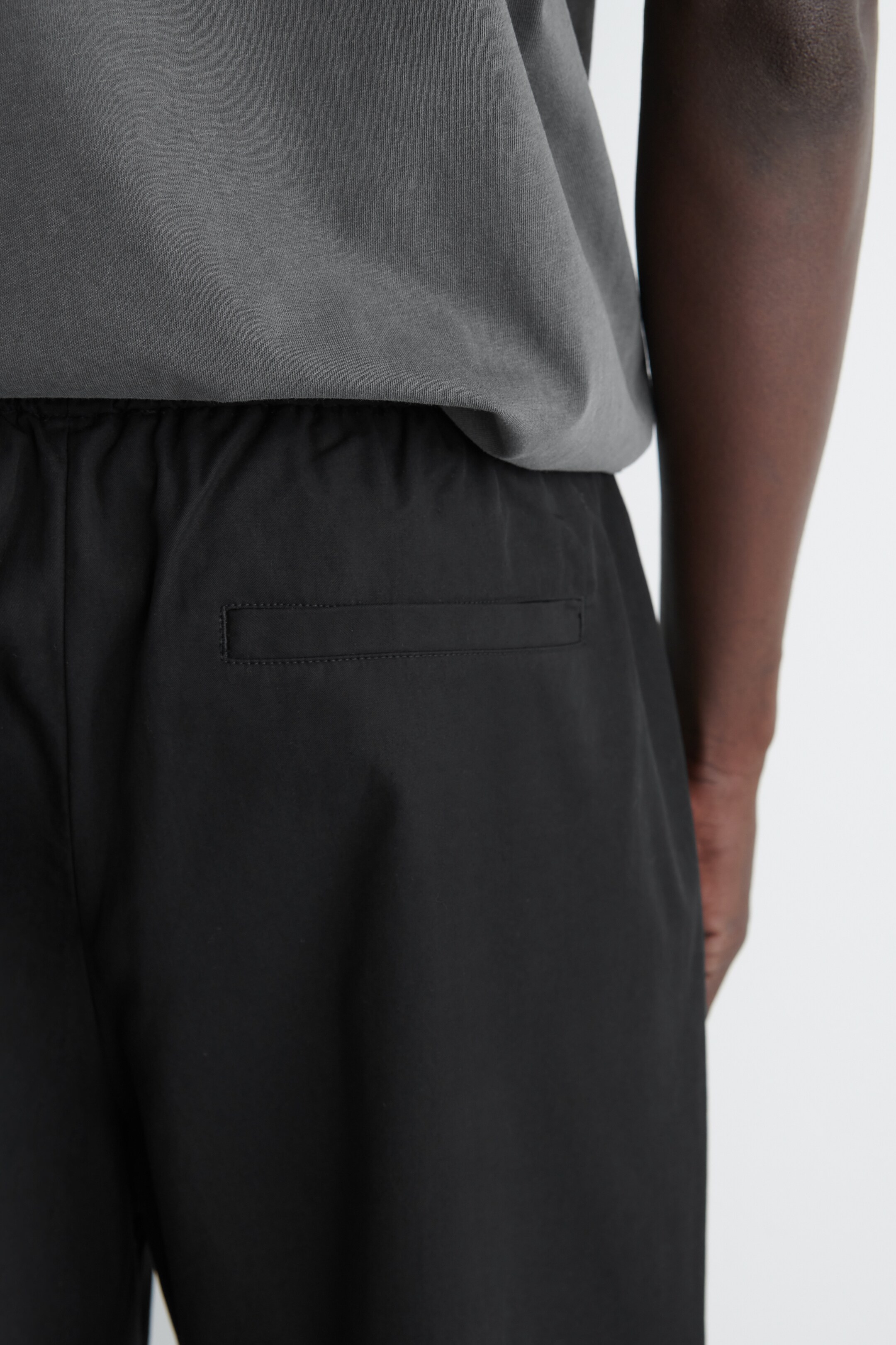 View larger image: Black joggers are being presented from a back view, featuring an elasticated waistband and a horizontal welt pocket on the back. The relaxed-fit joggers are appearing in a smooth black fabric, worn with a grey t-shirt partially visible above the waistband.