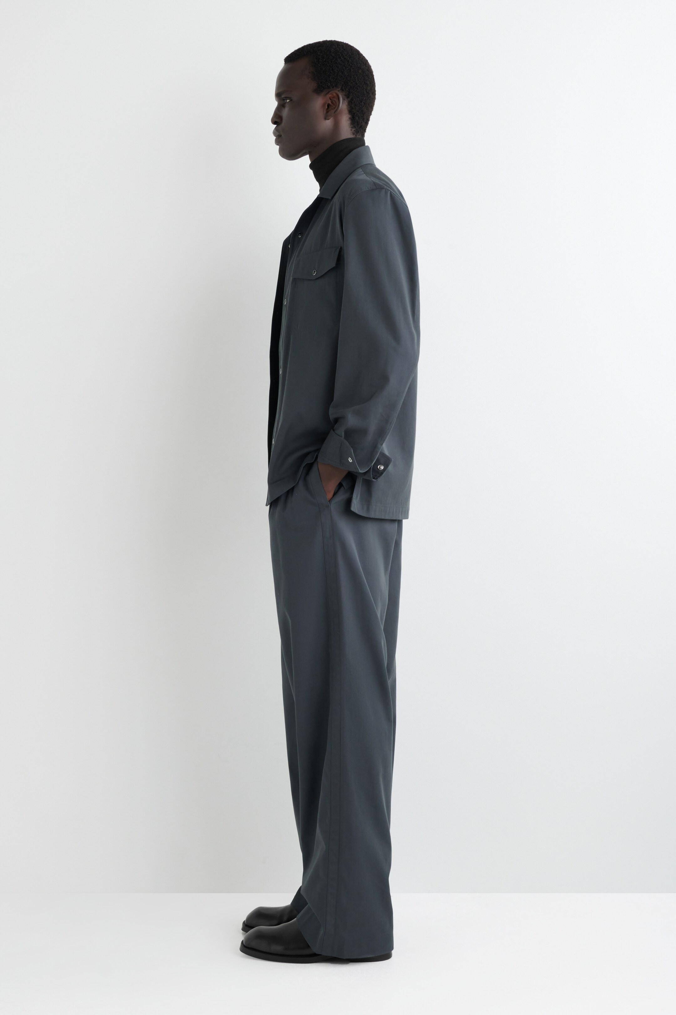 View larger image: A dark grey shirt jacket, featuring a collar, button-front closure, and two chest pockets, is being presented from a side profile. It is being worn over a black turtleneck and is paired with matching wide-leg trousers that are displaying a loose fit and are covering the tops of black shoes.