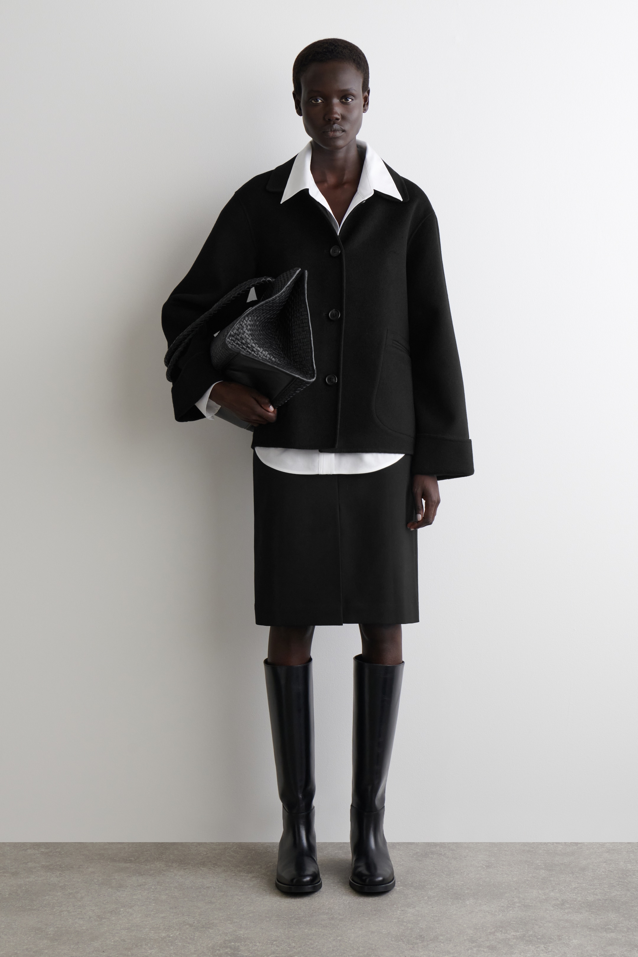 Agrandir l'image: Une veste noire à col, de coupe ample et carrée avec des manches larges est portée, avec une fermeture à trois boutons à l'avant et des poches plaquées arrondies. Elle est stylée par-dessus une chemise blanche, avec une jupe droite noire et des bottes noires jusqu'aux genoux. Un sac fourre-tout noir tissé est porté.