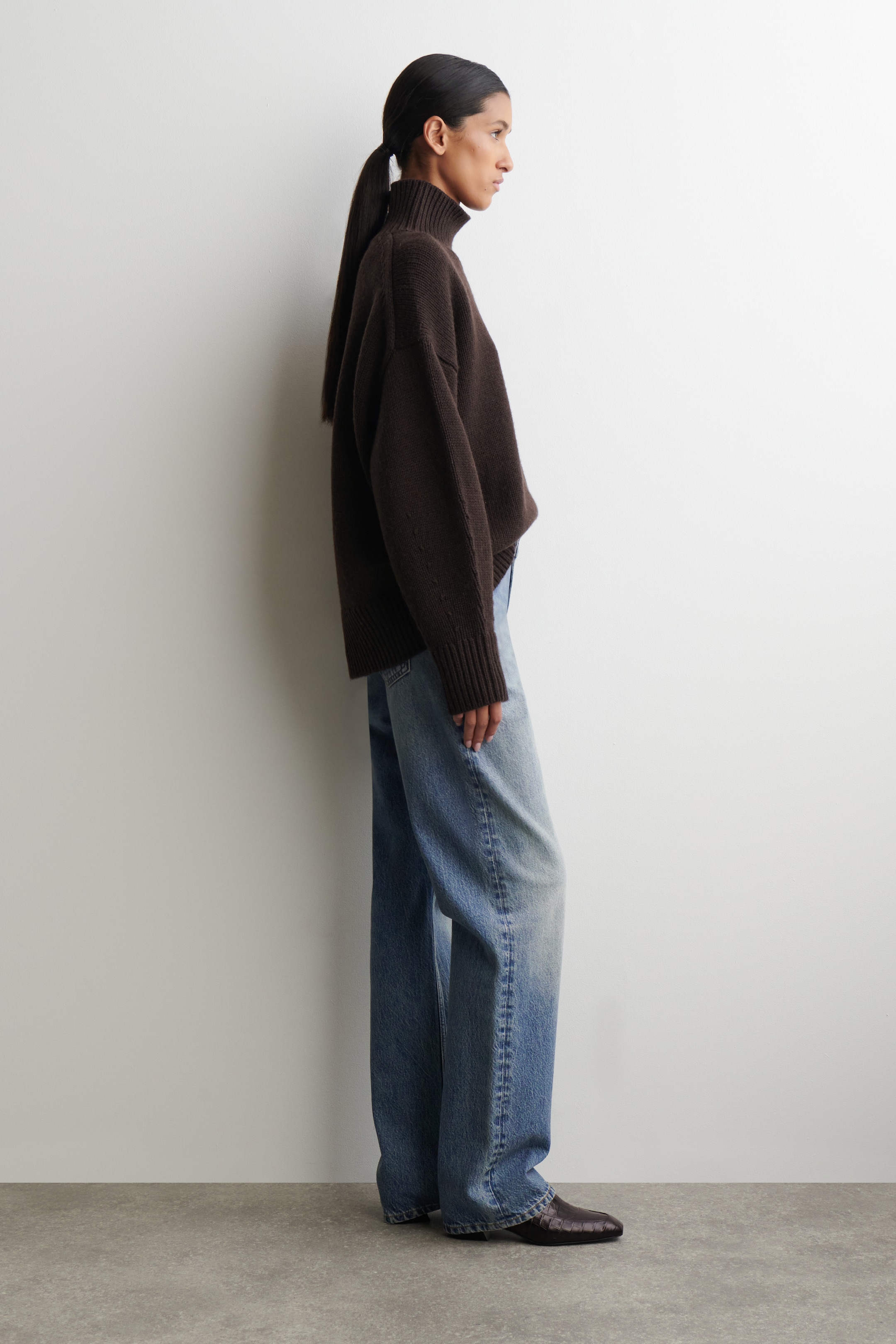 Agrandir l'image: Un pull à col roulé surdimensionné de couleur brun foncé est présenté de côté, avec un col roulé côtelé et des poignets côtelés. Il est porté par-dessus un jean en denim délavé clair.