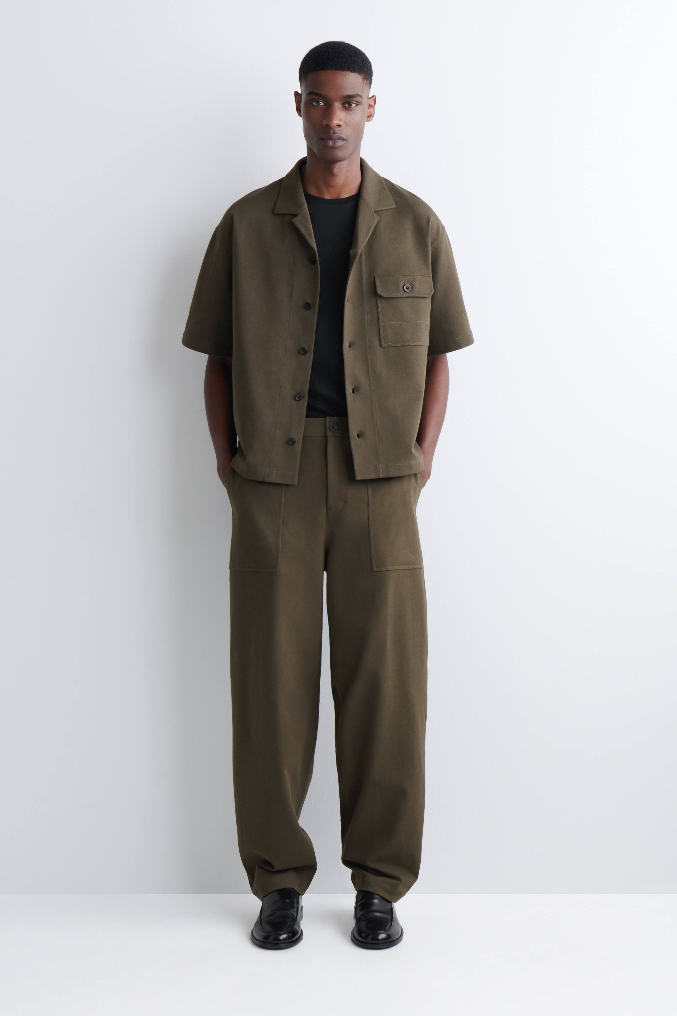 View larger image: An olive green co-ordinated set is being presented, facing forward, consisting of a short-sleeved shirt with a camp collar and a single flap pocket worn open over a black T-shirt, paired with wide-leg trousers featuring two large patch pockets.