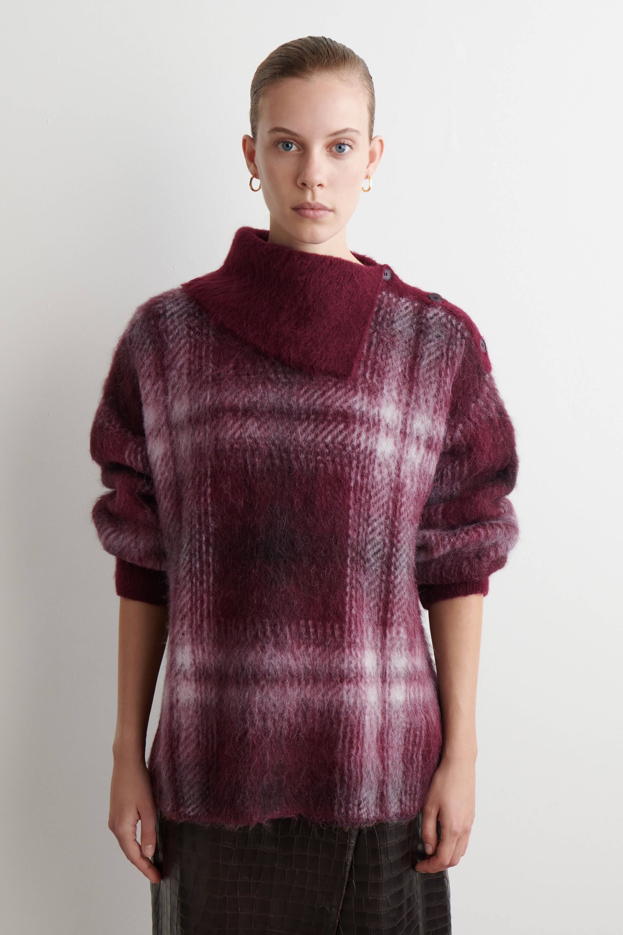 Agrandir l'image: Un pull en plaid duveteux à l'avant dans des tons de rouge, bordeaux et blanc est présenté. Il présente un col haut asymétrique plié avec des boutons visibles sur l'épaule droite, et des manches longues volumineuses se terminant par des poignets ajustés.