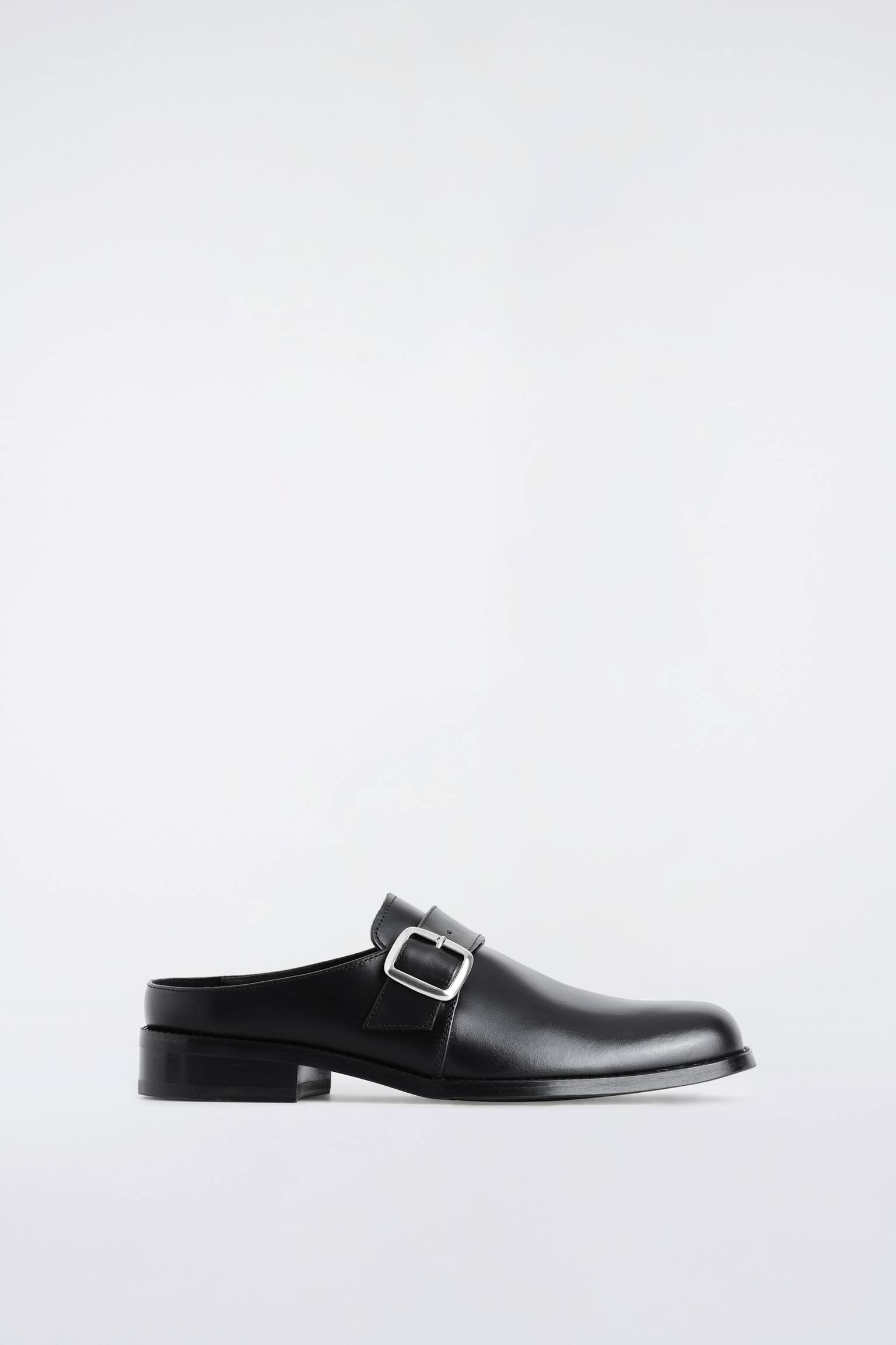 Un mule din piele neagrÄ este prezentat Ã®n profil lateral, orientat spre dreapta, evidenÈiind o parte superioarÄ din piele netedÄ, cu vÃ¢rf rotund Èi design cu cÄlcÃ¢i deschis. Pantoful are un toc jos, gros, Èi o bandÄ latÄ peste rist, fixatÄ cu o cataramÄ metalicÄ proeminentÄ, Ã®n ton argintiu.