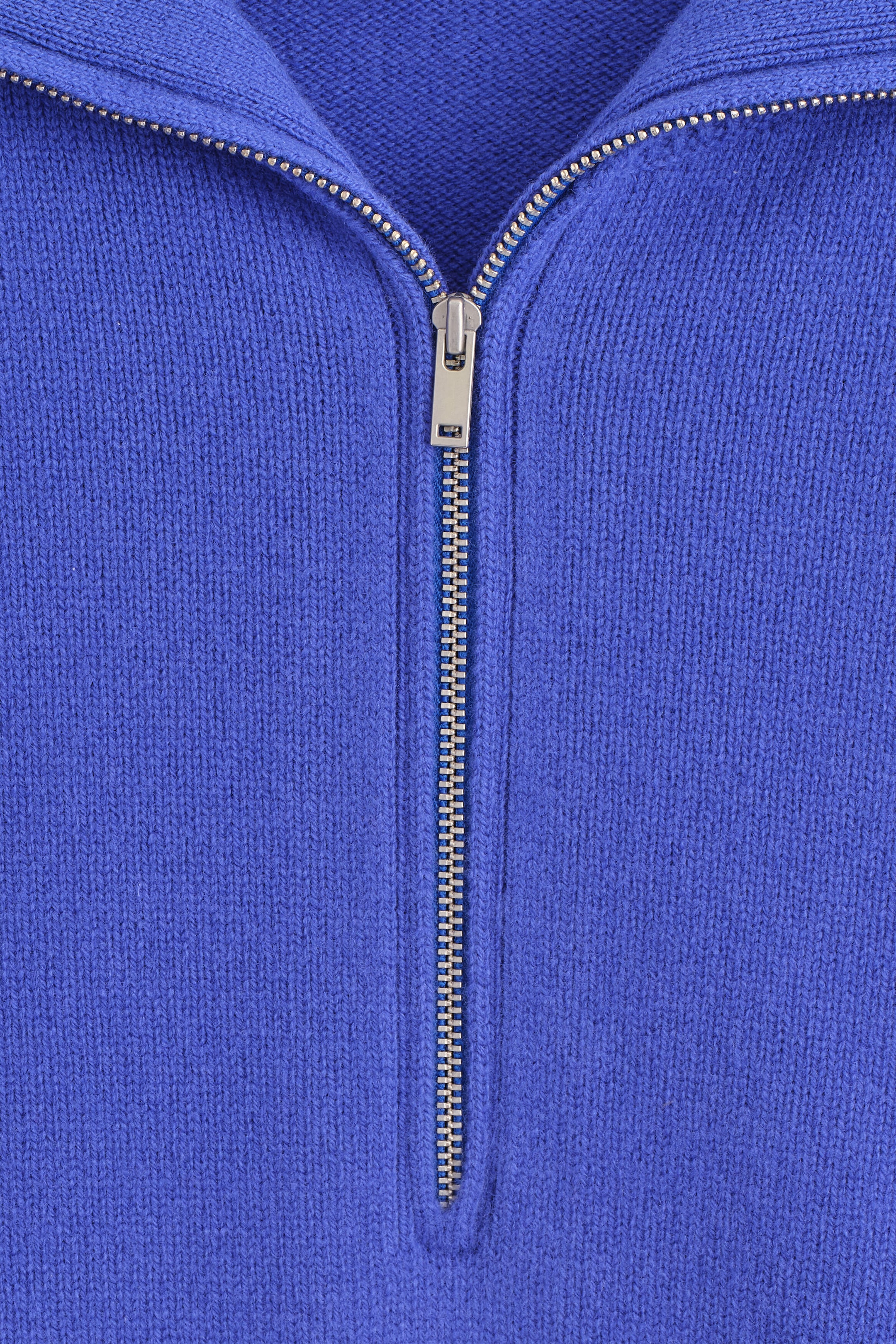 A close-up front view of a blue knitted sweater featuring a silver-tone metal zipper partially unzipped to create a wide collar, highlighting the fine-knit texture of the fabric. A close-up front view of a blue knitted sweater featuring a silver-tone metal zipper partially unzipped to create a wide collar, highlighting the fine-knit texture of the fabric.
