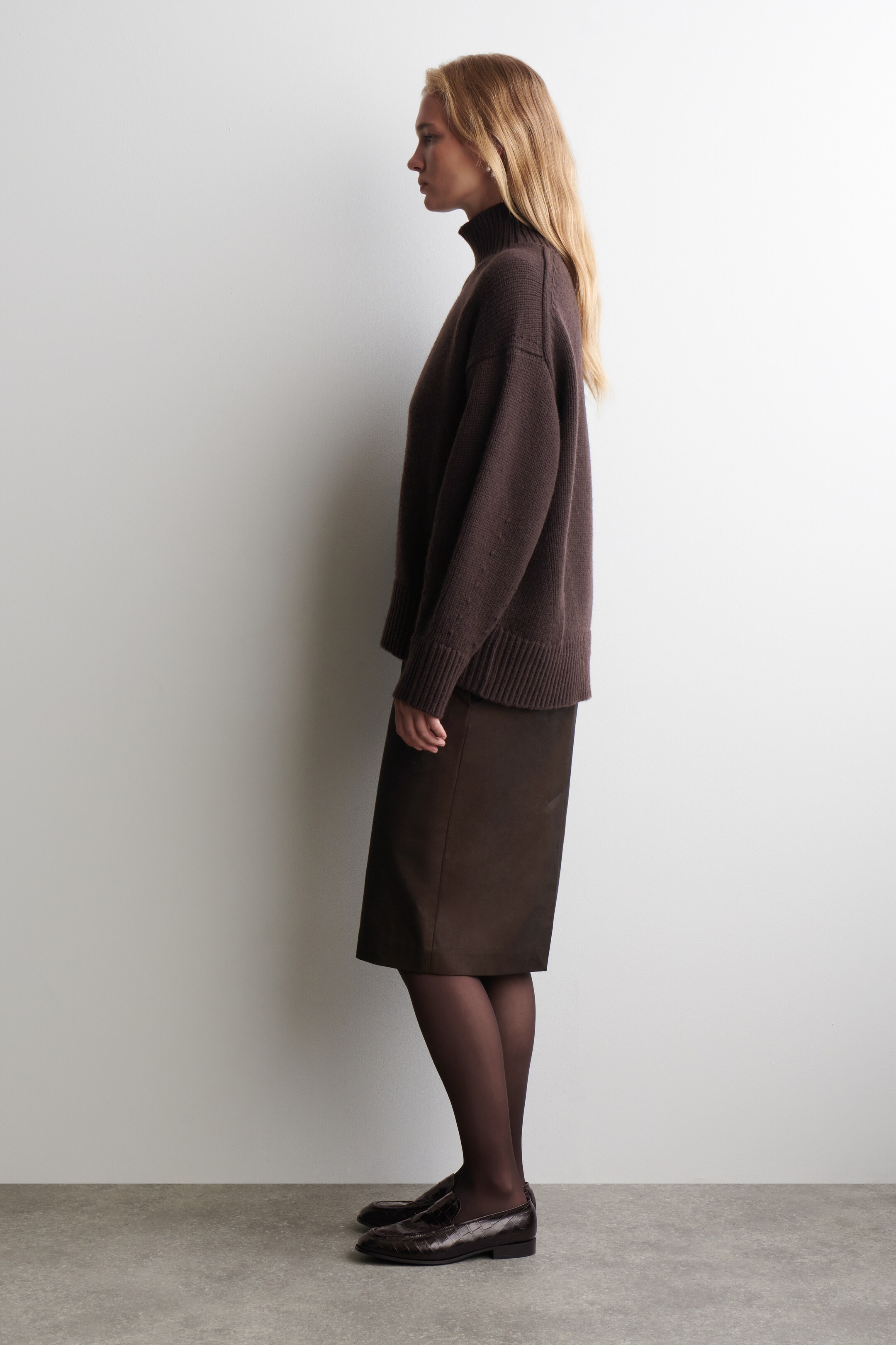 View larger image: A dark brown relaxed-fit turtleneck sweater with ribbed cuffs and hem is being presented from a side profile. It is being styled with a dark brown straight knee-length skirt and dark brown textured loafers.