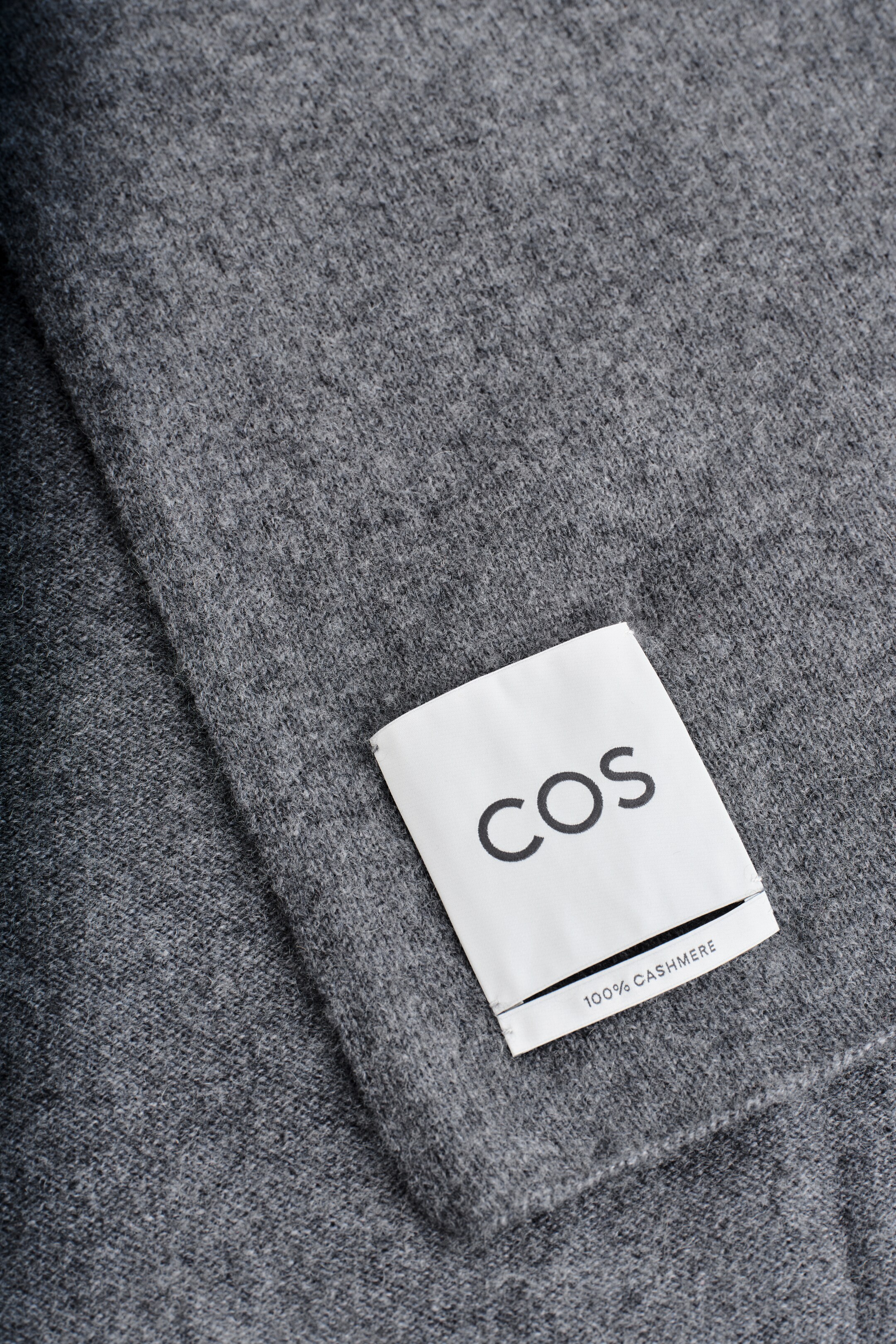 View larger image: A grey cashmere scarf is being shown in a close-up, highlighting its soft, finely knit texture. A white label, bearing the "COS" logo and "100% CASHMERE," is resting on the scarf's surface.