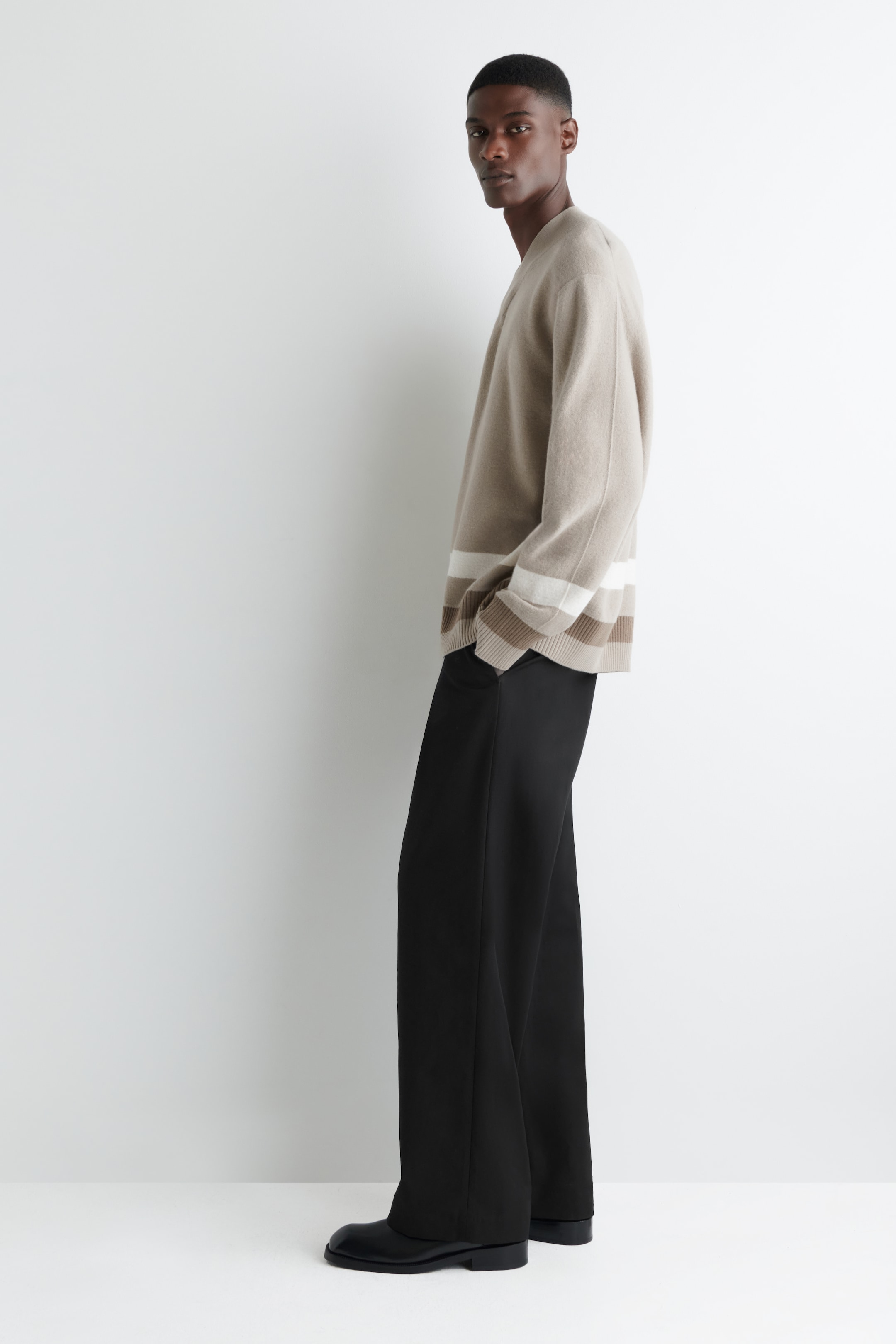 View larger image: A light brown V-neck sweater, featuring white and brown horizontal stripes along the hem, is being worn with black wide-leg trousers and black dress shoes. The ensemble is being presented from a side angle, highlighting the relaxed fit of the sweater and the fluid drape of the trousers.