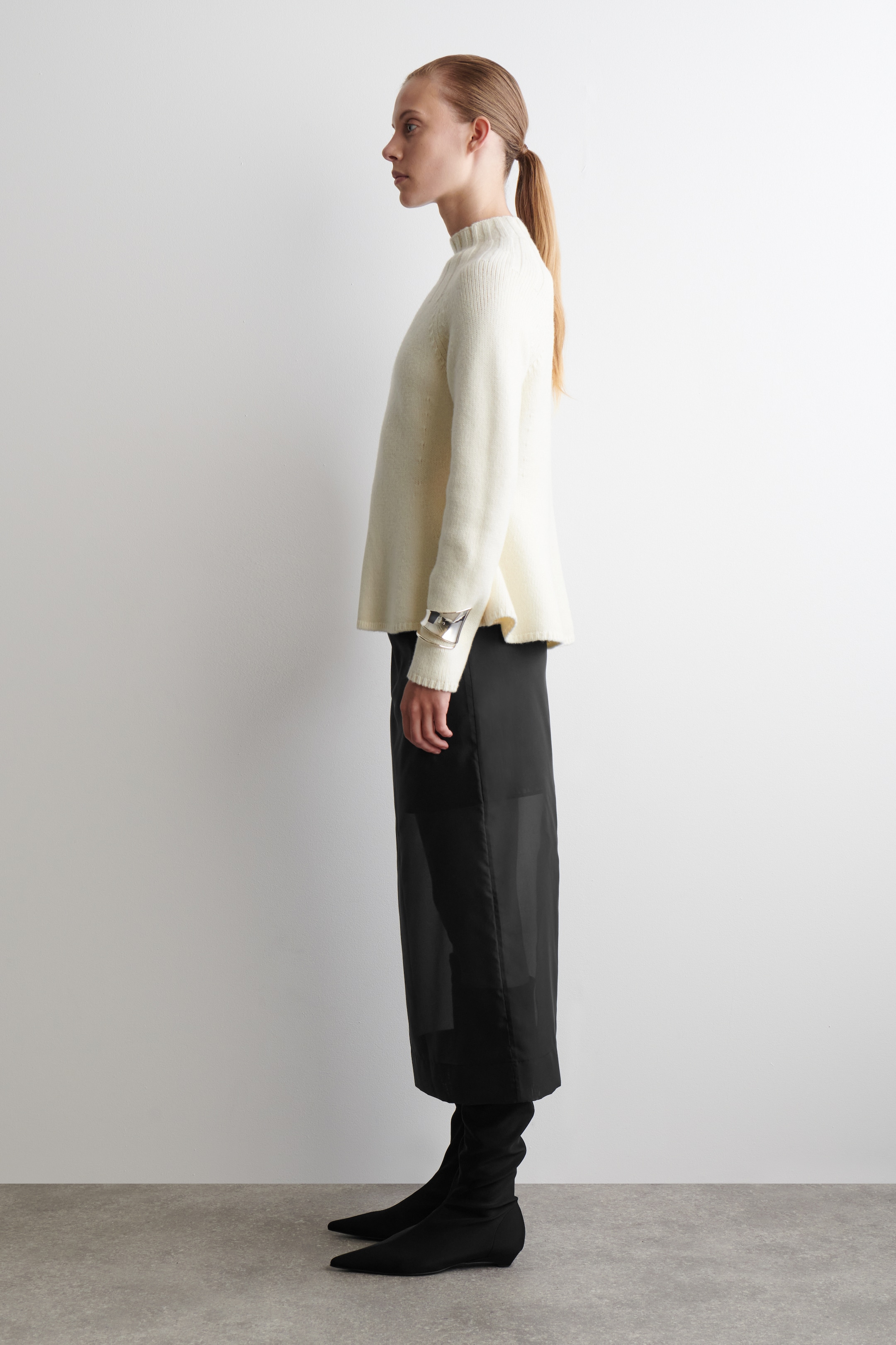 View larger image: A person is standing in profile, wearing a cream sweater featuring a ribbed high neck, long sleeves, and a subtly flared hem. They are also wearing a black midi skirt with sheer panel details and black pointed-toe boots with slouching shafts.