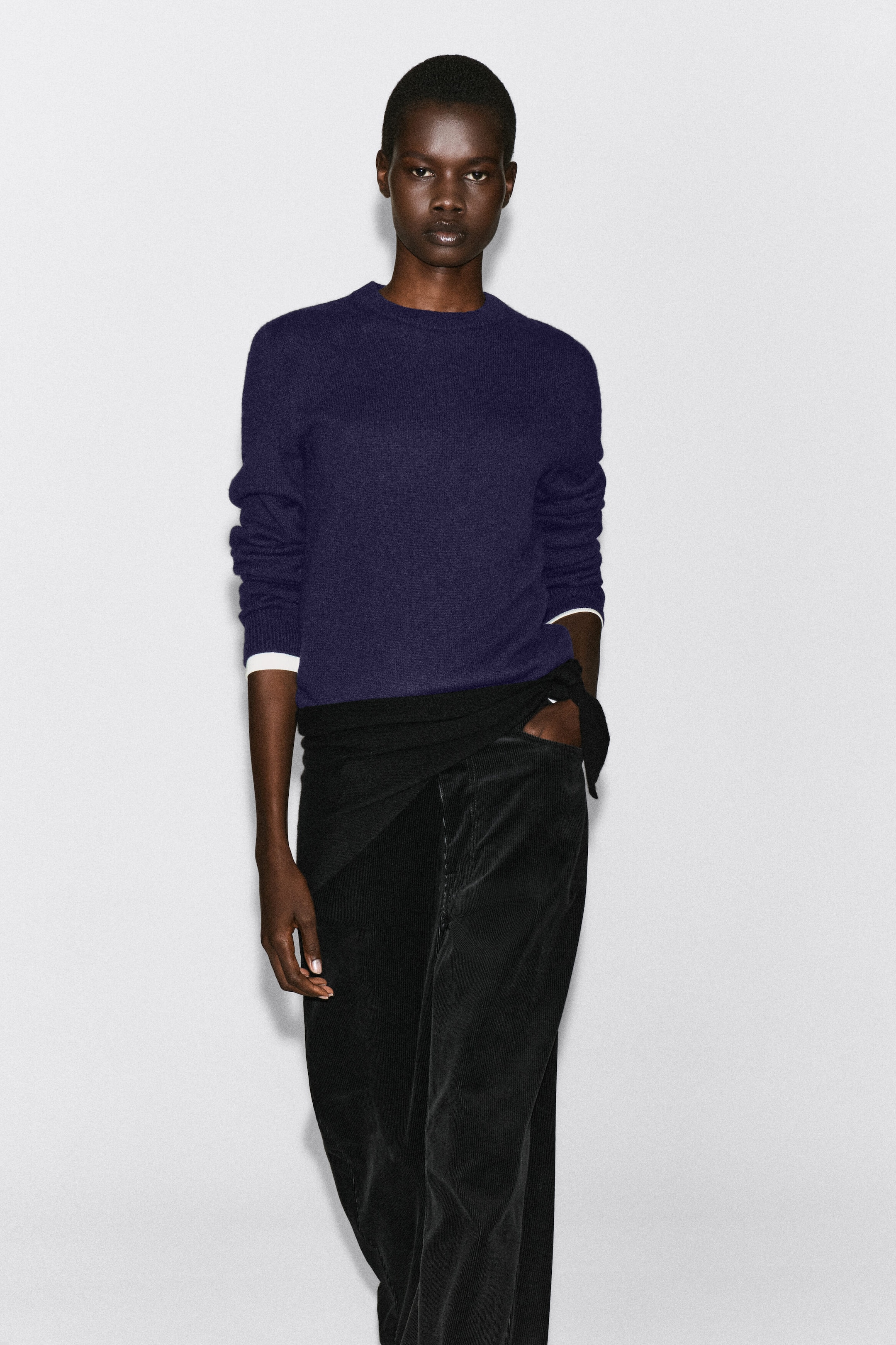 Un pull en maille col rond violet foncé est porté par le mannequin, superposé à une chemise blanche qui dépasse aux poignets. Le pull est associé à un vêtement noir noué à la taille et à un pantalon en velours côtelé noir. Un pull en maille col rond violet foncé est porté par le mannequin, superposé à une chemise blanche qui dépasse aux poignets. Le pull est associé à un vêtement noir noué à la taille et à un pantalon en velours côtelé noir.