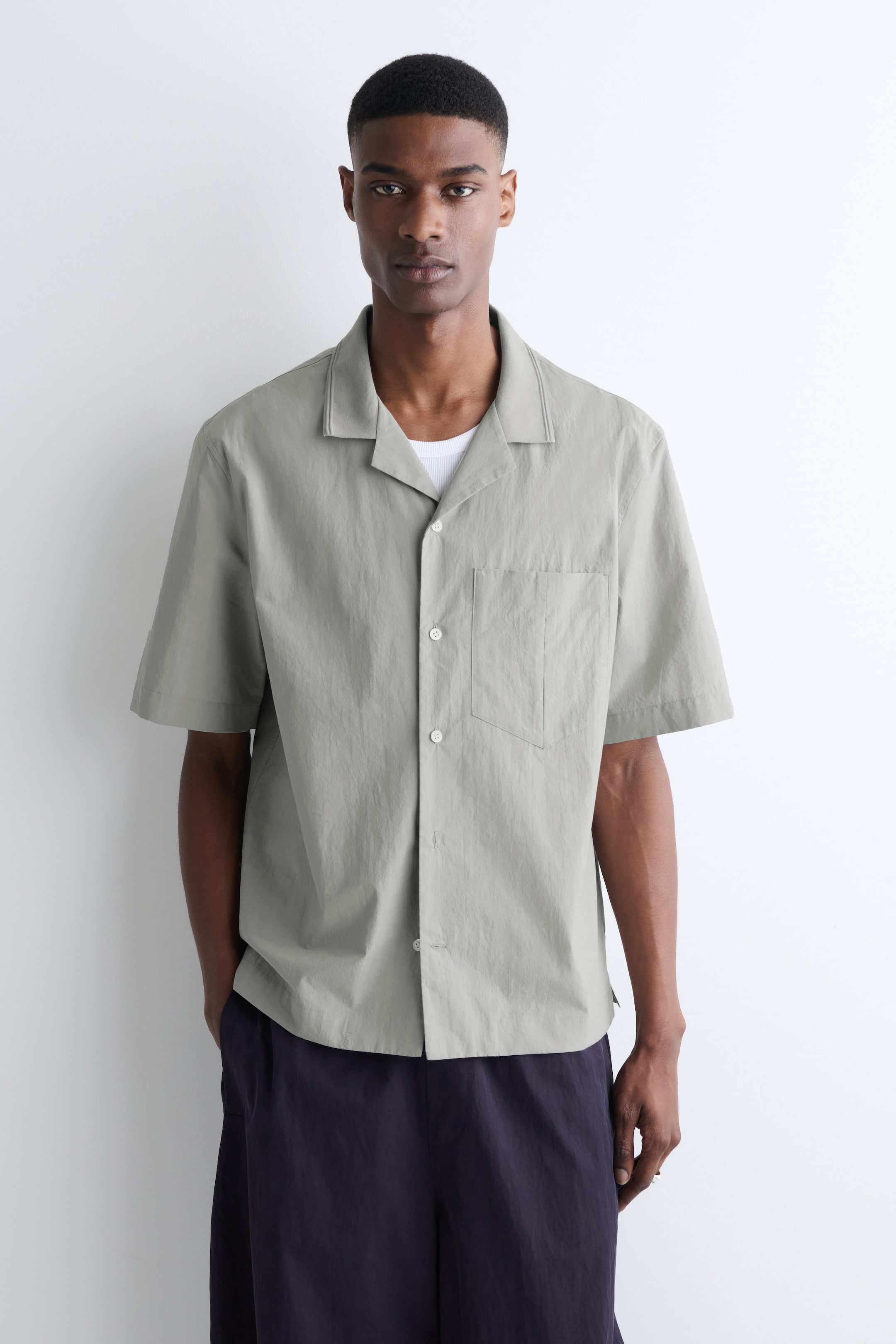 Größeres Bild anzeigen: Ein hellgraues Kurzarmhemd mit Reverskragen und einer aufgesetzten Brusttasche auf der linken Seite. Es präsentiert einen Knopfverschluss mit weißen Knöpfen und wird offen über einem weißen, gerippten Unterhemd getragen.