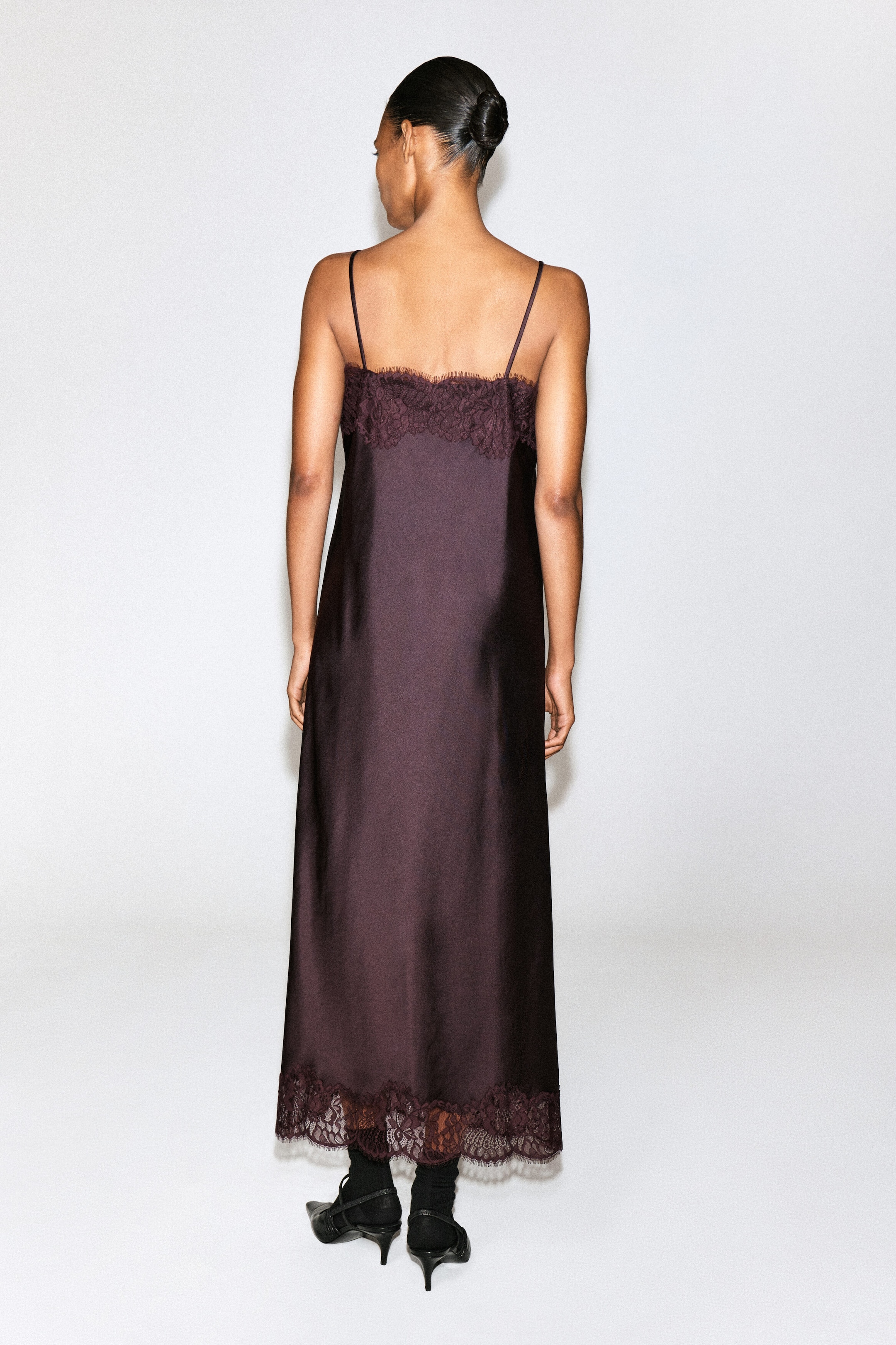 Ein aus Satin gefertigtes Slipkleid mit Spitzensaum in dunklem Pflaumenton wird von dem Model getragen, das mit dem Rücken zur Kamera steht. Das knöchellange Kleid verfügt über schmale Spaghettiträger, eine zarte Spitzenverzierung am oberen Rücken und eine passende Spitzenverzierung am Saum.