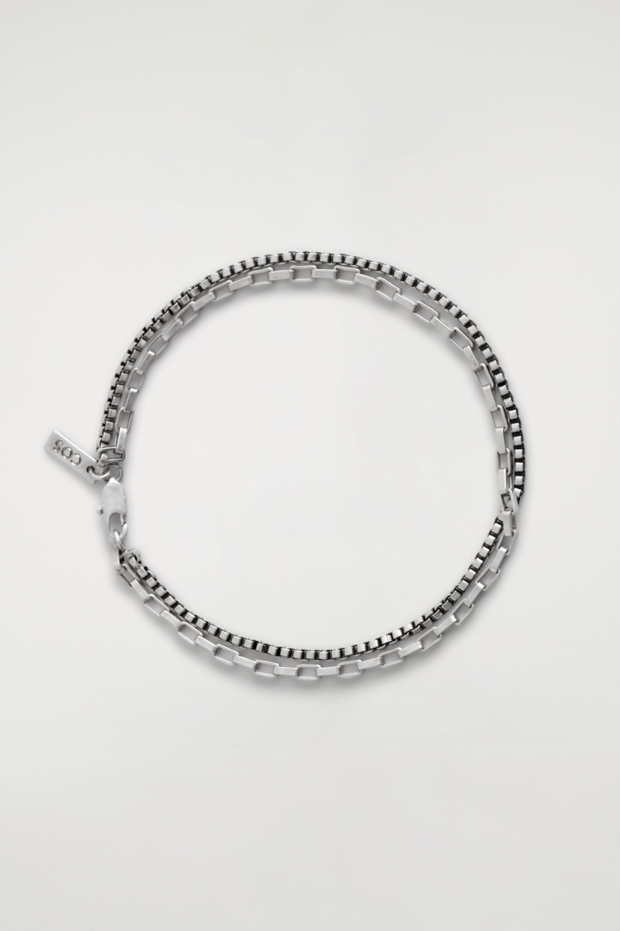 Grösseres Bild ansehen: Ein doppelreihiges Armband zeigt zwei unterschiedliche silberfarbene Ketten: eine hellere rechteckige Gliederkette und eine dunklere Boxkette. Die Ketten sind zusammen geschichtet und bilden eine kreisförmige Präsentation, mit einem sichtbaren Karabinerverschluss und einem kleinen rechteckigen Anhänger, der mit "COS" graviert ist.