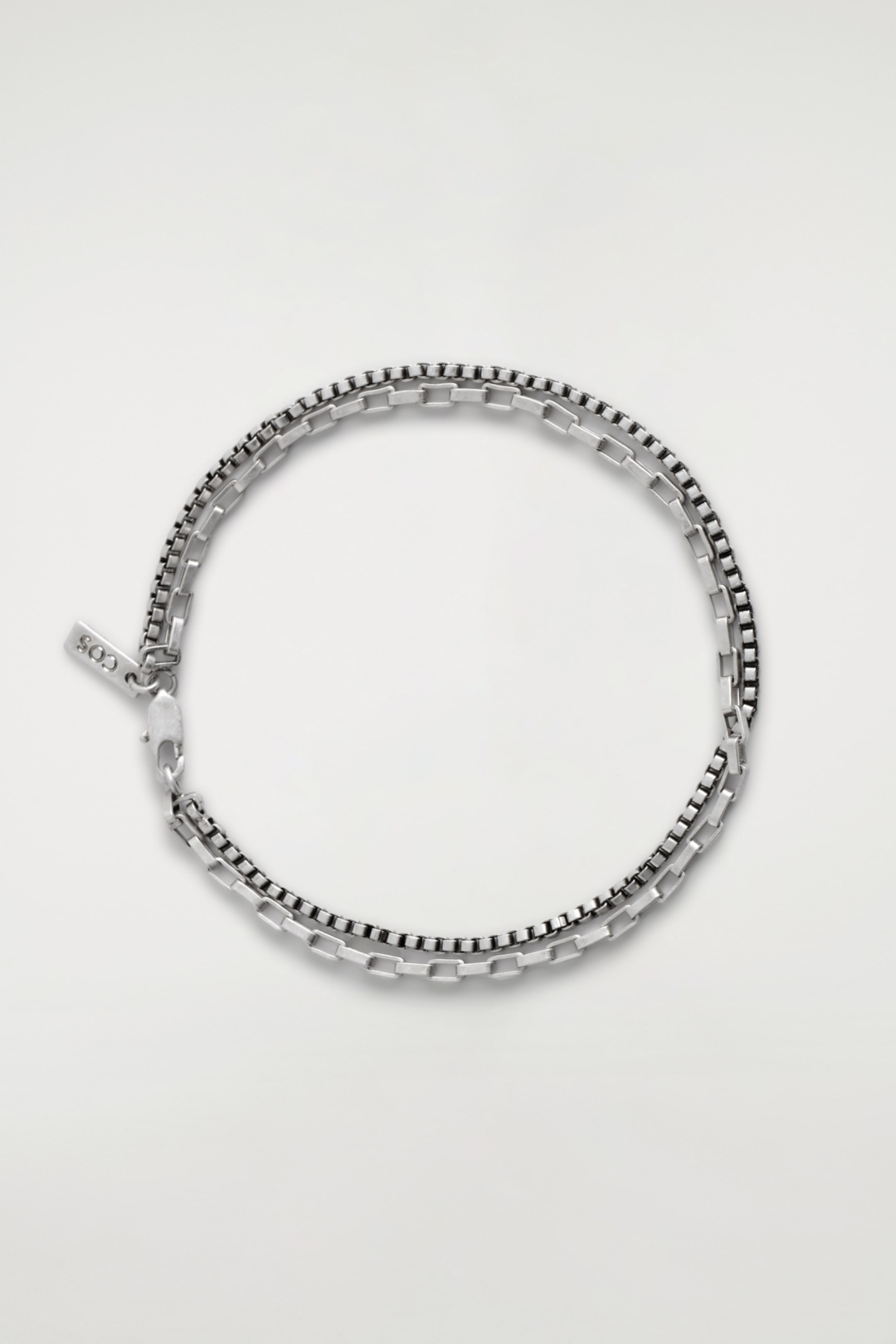 Grotere afbeelding bekijken: Een dubbelgelaagde armband toont twee verschillende zilverkleurige kettingen: een helderdere rechthoekige schakelketting en een donkerdere boxketting. De kettingen zijn samen gelaagd, vormen een cirkelvormige presentatie, met een zichtbare karabijnsluiting en een klein rechthoekig label gegraveerd met "COS".