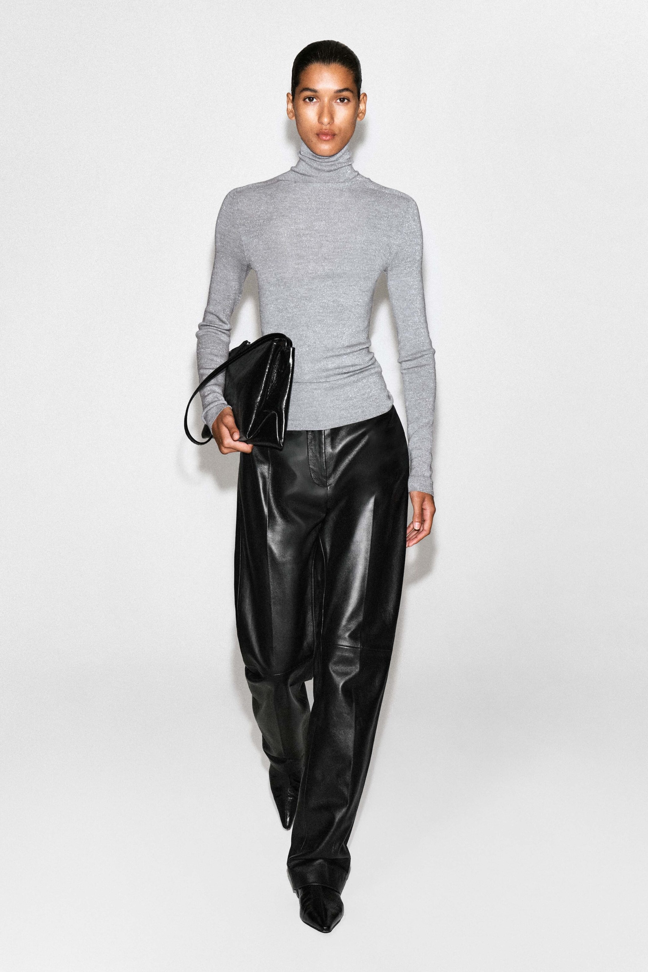 The sparkly seamless wool funnel-neck top is shown from the front in a slim-fit silhouette, crafted from a light gray metallic knit. It features a high, snug funnel neck and long sleeves, styled untucked over black leather trousers to highlight its clean, seamless hem.