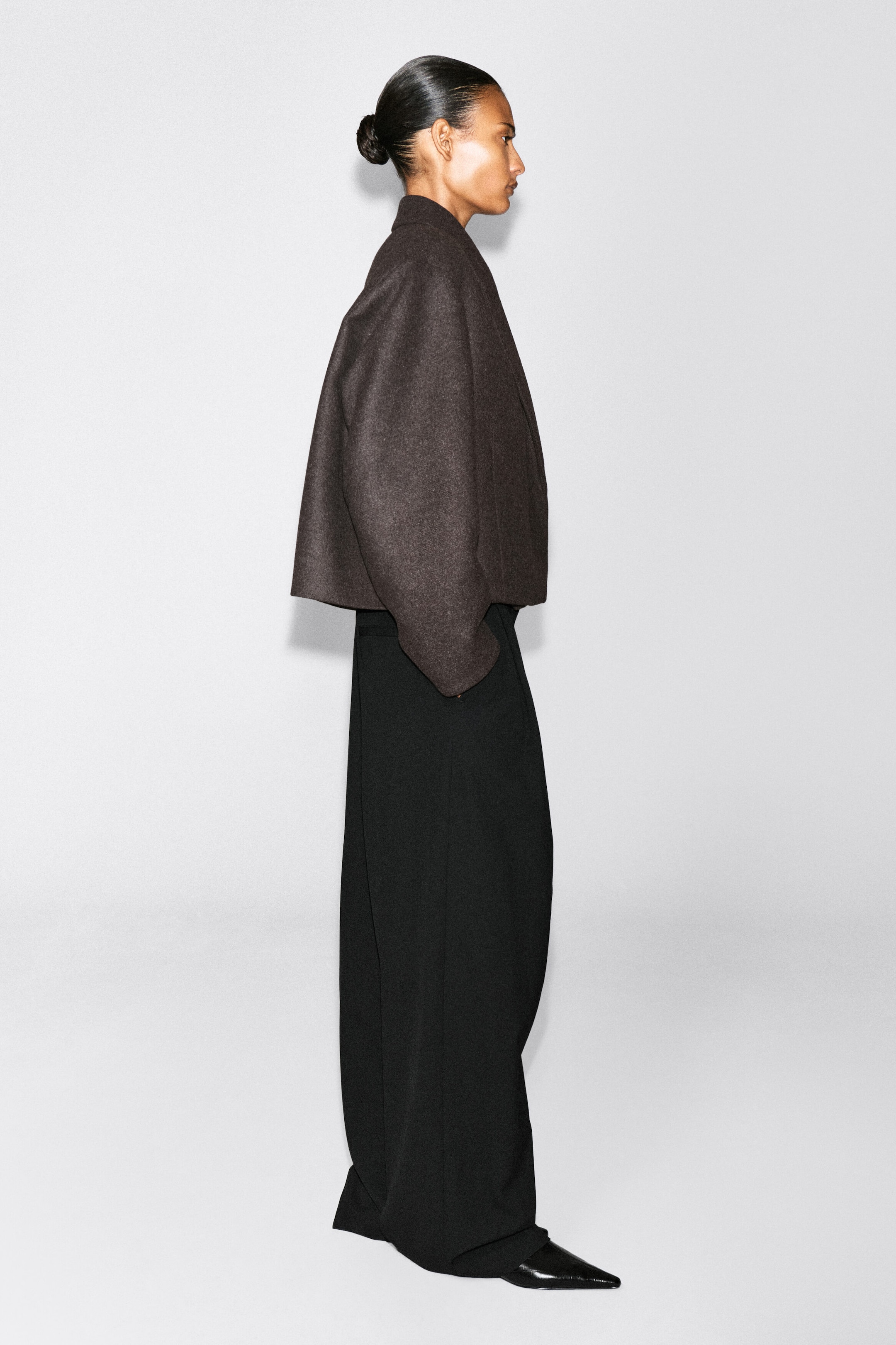 An oversized, cropped boiled-wool blend blazer in dark brown is being worn by the model, who is standing in profile. The blazer features a structured, voluminous silhouette with curved sleeves and is styled with black wide-leg trousers. An oversized, cropped boiled-wool blend blazer in dark brown is being worn by the model, who is standing in profile. The blazer features a structured, voluminous silhouette with curved sleeves and is styled with black wide-leg trousers.