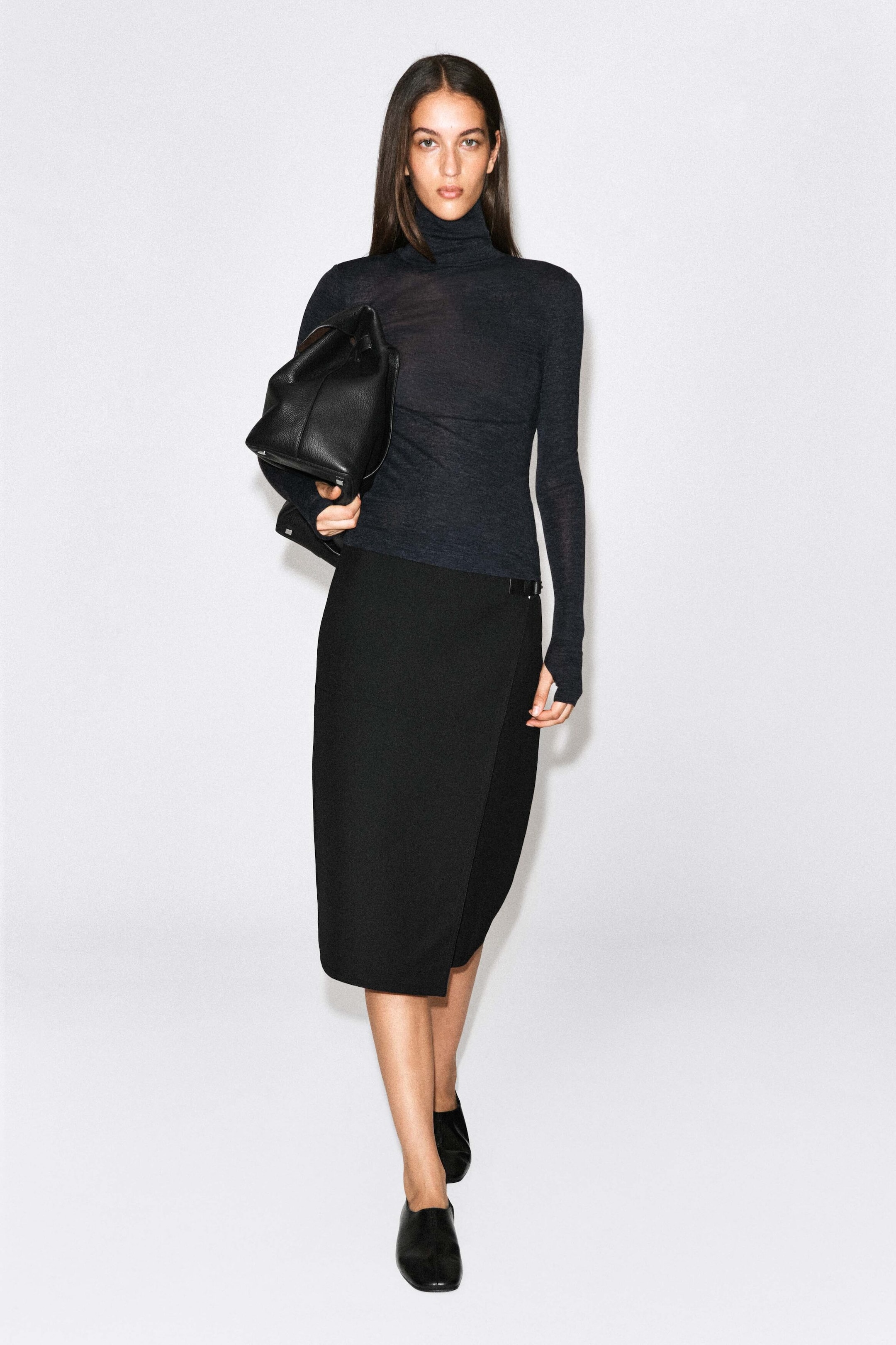 The model is wearing a charcoal gray merino wool high-neck top featuring a fitted silhouette, semi-sheer texture, and long sleeves. The top is paired with a black knee-length wrap skirt and a black leather bag carried under the arm. The model is wearing a charcoal gray merino wool high-neck top featuring a fitted silhouette, semi-sheer texture, and long sleeves. The top is paired with a black knee-length wrap skirt and a black leather bag carried under the arm.