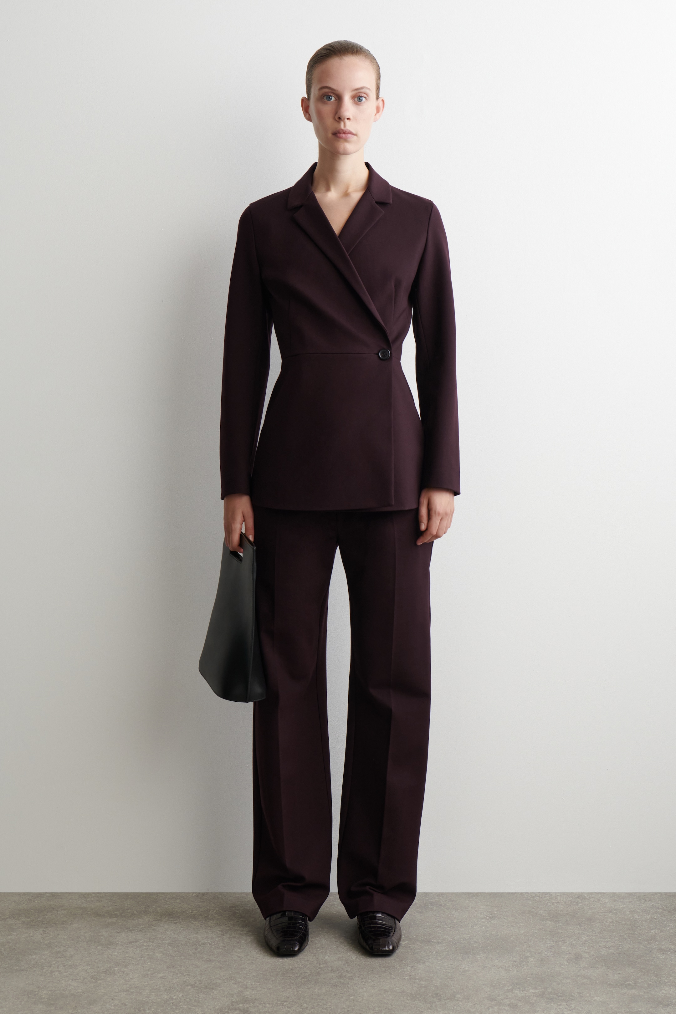 Visualizza immagine più grande: Un completo da donna color prugna scuro è esposto da una prospettiva frontale, caratterizzato da un blazer su misura con revers a tacca e chiusura a un bottone, abbinato a pantaloni a gamba larga. Una borsa strutturata nera è tenuta in mano e sono visibili scarpe basse scure, completando l'insieme.