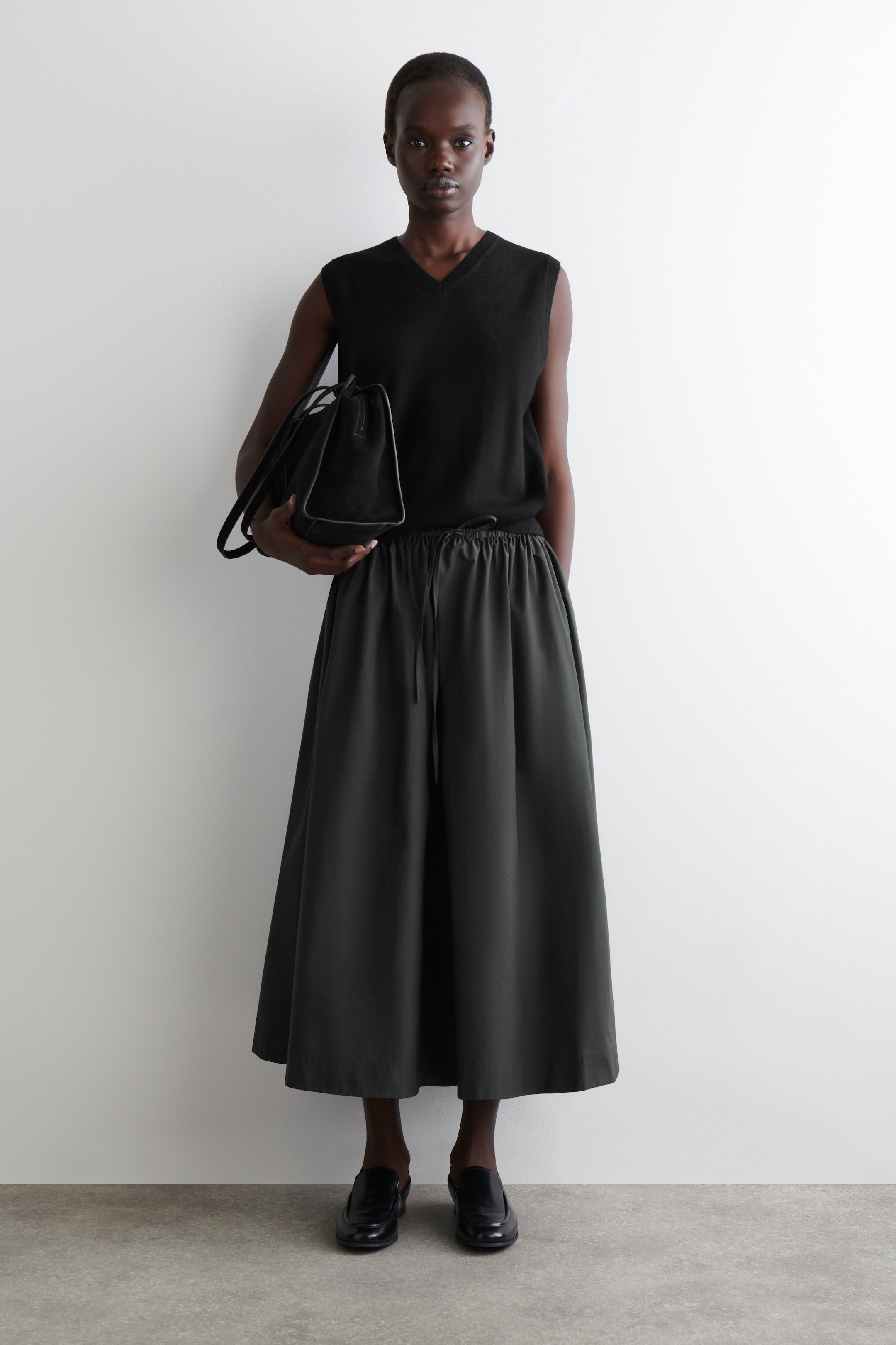 Agrandir l'image: Un gilet en tricot noir sans manches à col en V est assorti à une jupe midi à jambes larges en charbon foncé avec une taille à cordon visible. Un sac fourre-tout structuré noir est tenu, et des mocassins noirs à enfiler sont portés.