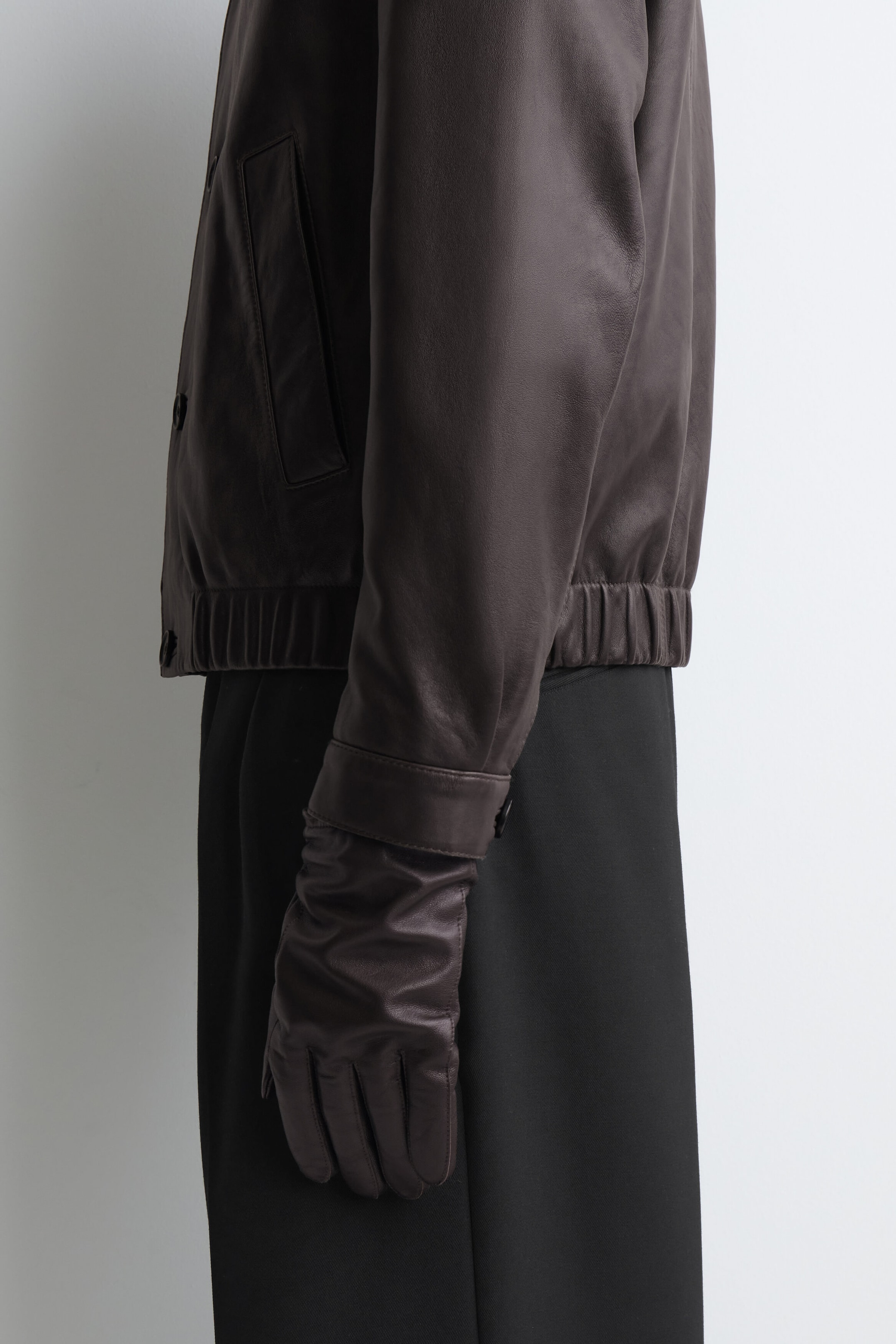 Dark brown leather gloves featuring topstitched details and a buttoned cuff are being worn by the model, layered under the sleeve of a matching dark brown leather jacket. Dark brown leather gloves featuring topstitched details and a buttoned cuff are being worn by the model, layered under the sleeve of a matching dark brown leather jacket.