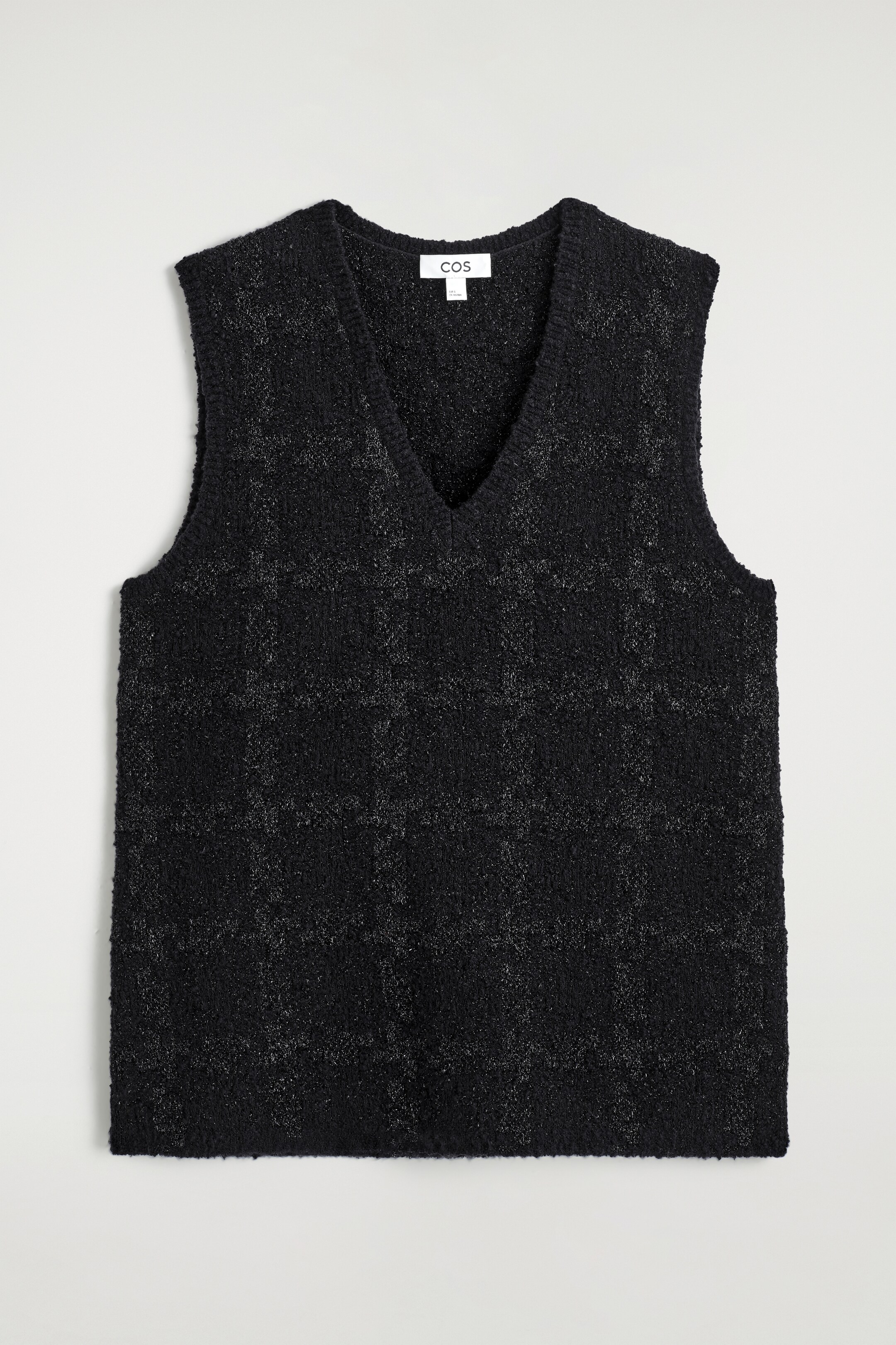 Agrandir l'image: Un gilet noir à col en V est présenté de face, avec un tricot bouclé tactile. Un motif à carreaux subtil est visible sur le gilet, formé par des fils argentés qui captent la lumière.