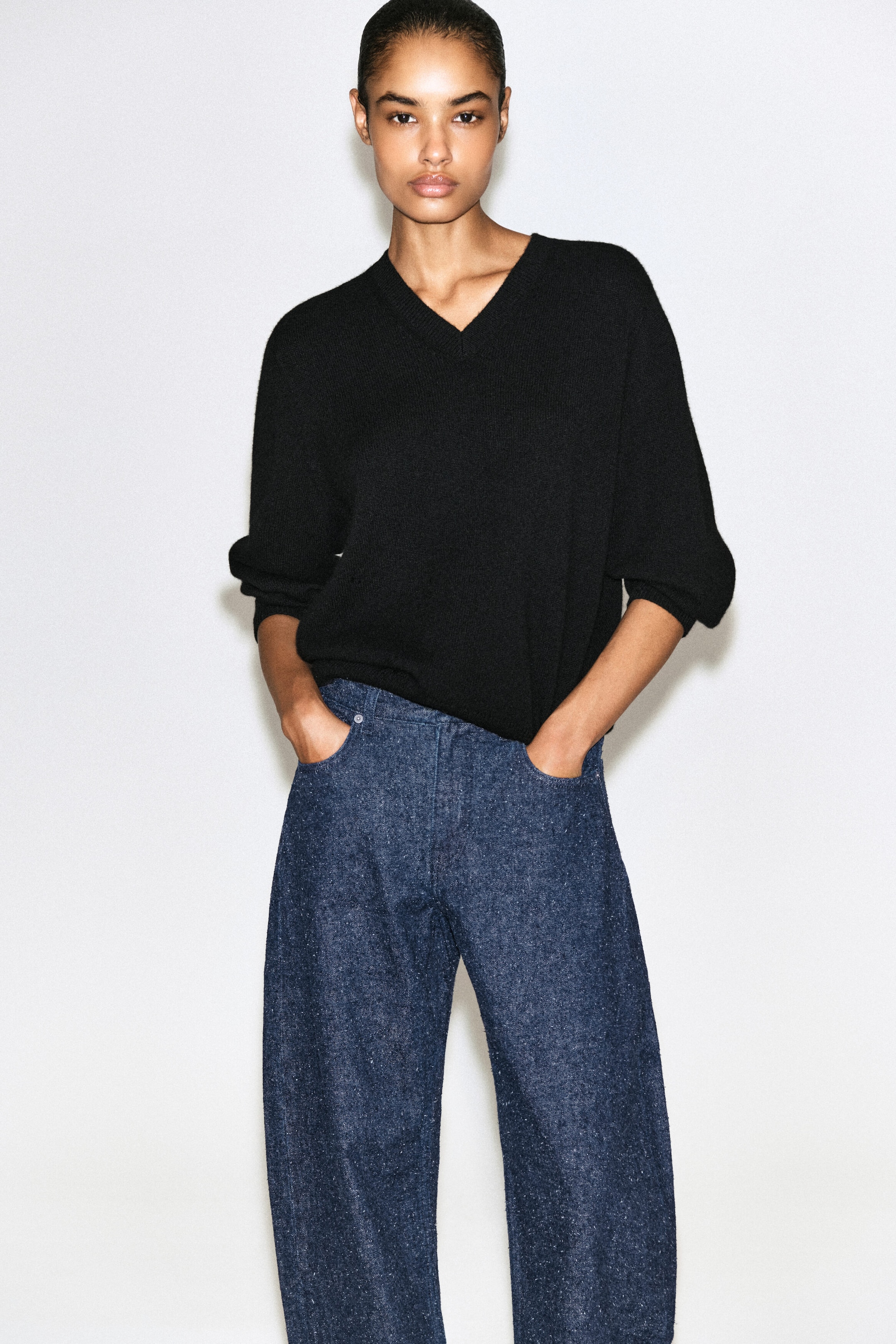 A black knit V-neck sweater with a relaxed fit and ribbed neckline is shown from the front, paired with dark blue textured wide-leg denim jeans. The sweater is styled with pushed-up sleeves, and the model is standing with their hands resting in the front pockets of the high-waisted jeans. A black knit V-neck sweater with a relaxed fit and ribbed neckline is shown from the front, paired with dark blue textured wide-leg denim jeans. The sweater is styled with pushed-up sleeves, and the model is standing with their hands resting in the front pockets of the high-waisted jeans.