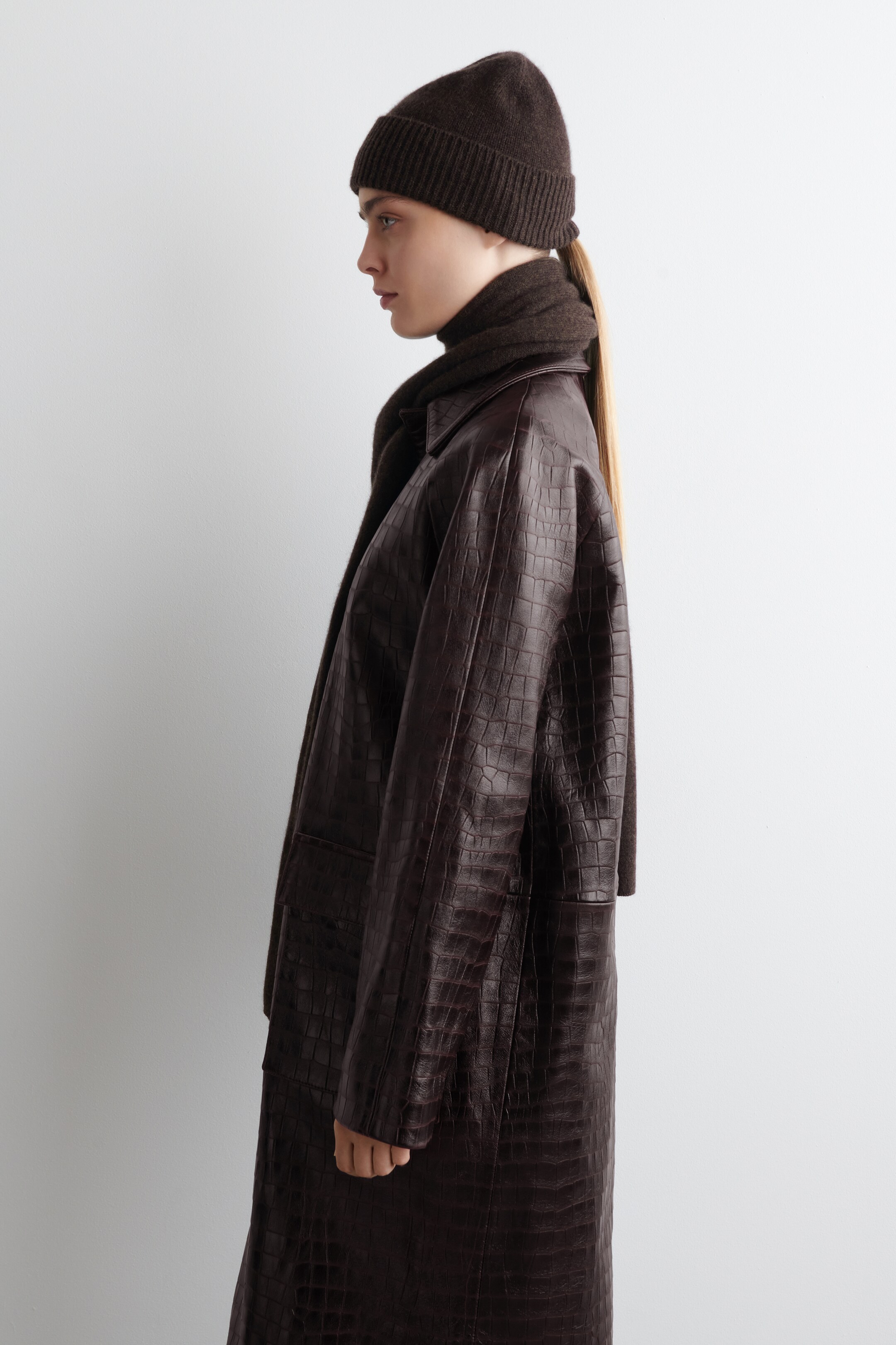 Agrandir l'image: Un manteau marron foncé, embossé crocodile et une jupe assortie sont présentés de profil. Le manteau est doté de manches longues et d'un col structuré, tandis qu'un bonnet côtelé marron foncé couvre la tête, et une écharpe douce marron foncé est enroulée autour du cou.