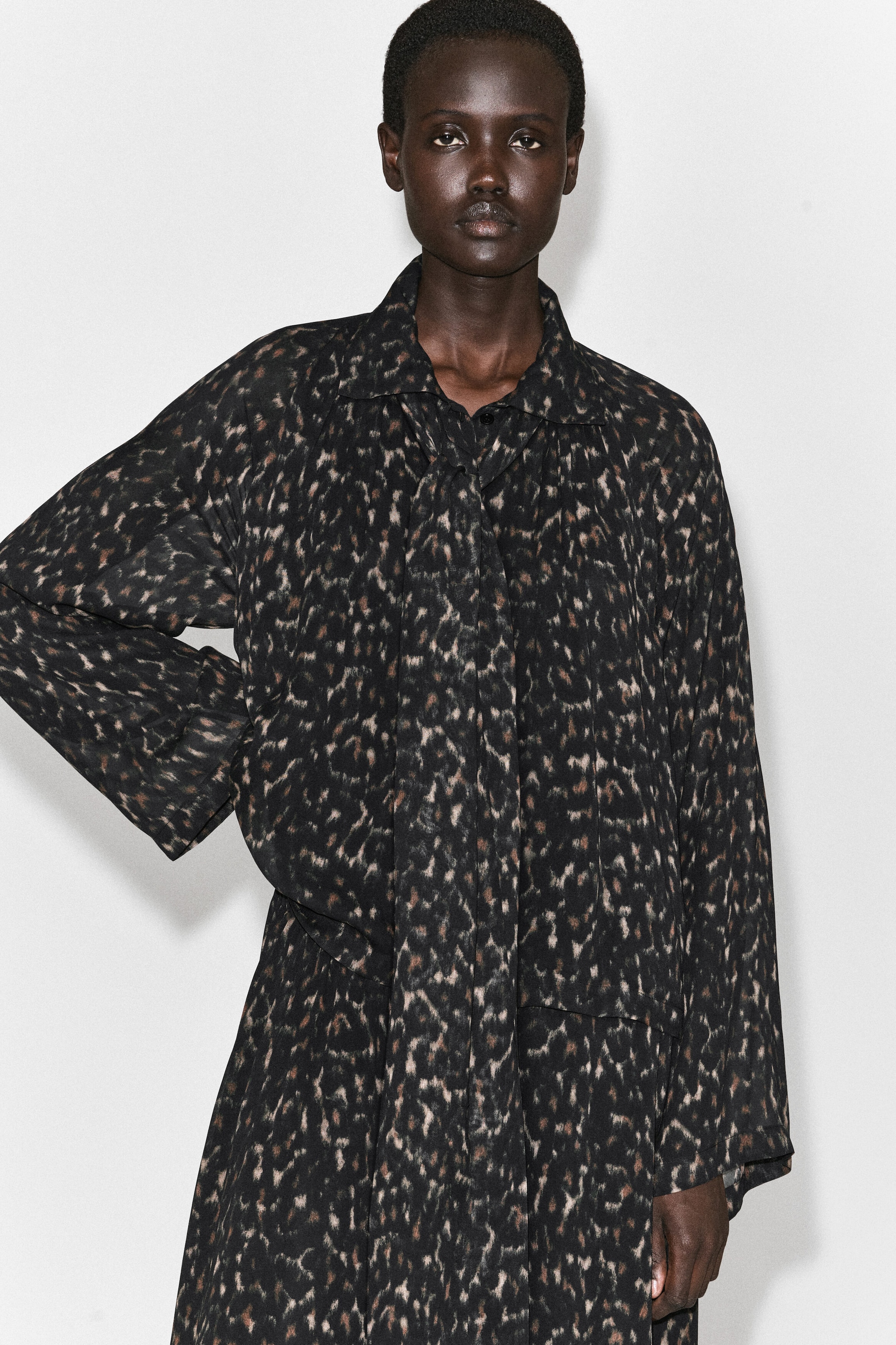 The leopard-print fluid dress with scarf detail showcases a dark, abstract animal-print pattern, a pointed collar, and a matching draped scarf detail hanging down the centre front. The dress features a relaxed fit with long, wide-cut sleeves. The leopard-print fluid dress with scarf detail showcases a dark, abstract animal-print pattern, a pointed collar, and a matching draped scarf detail hanging down the centre front. The dress features a relaxed fit with long, wide-cut sleeves.