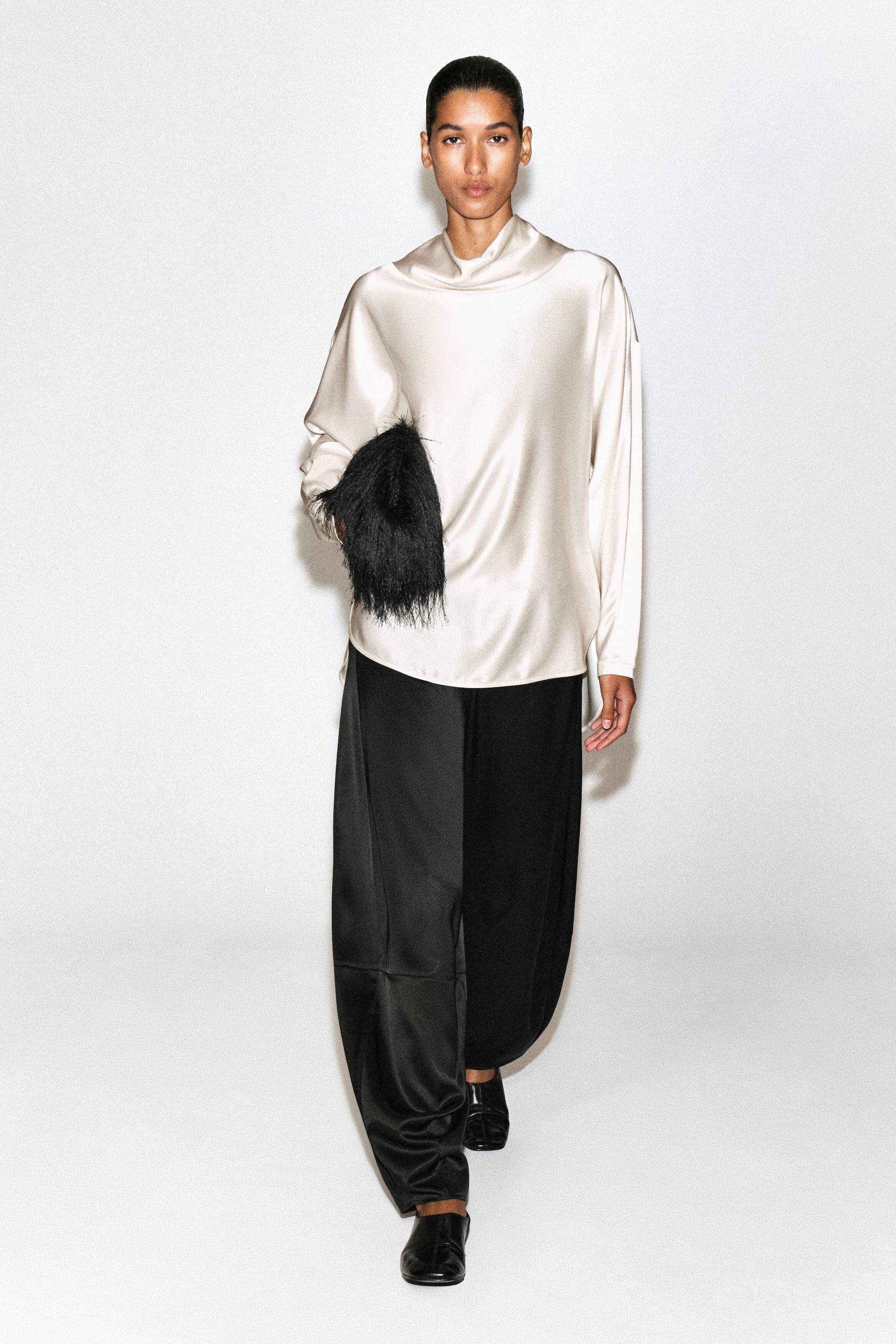 A cream-colored cowl-neck satin blouse is shown from the front, featuring a draped cowl neckline, a relaxed fit, and long sleeves. The blouse hangs loosely over wide-leg black trousers, highlighting its smooth, lustrous texture and fluid drape.