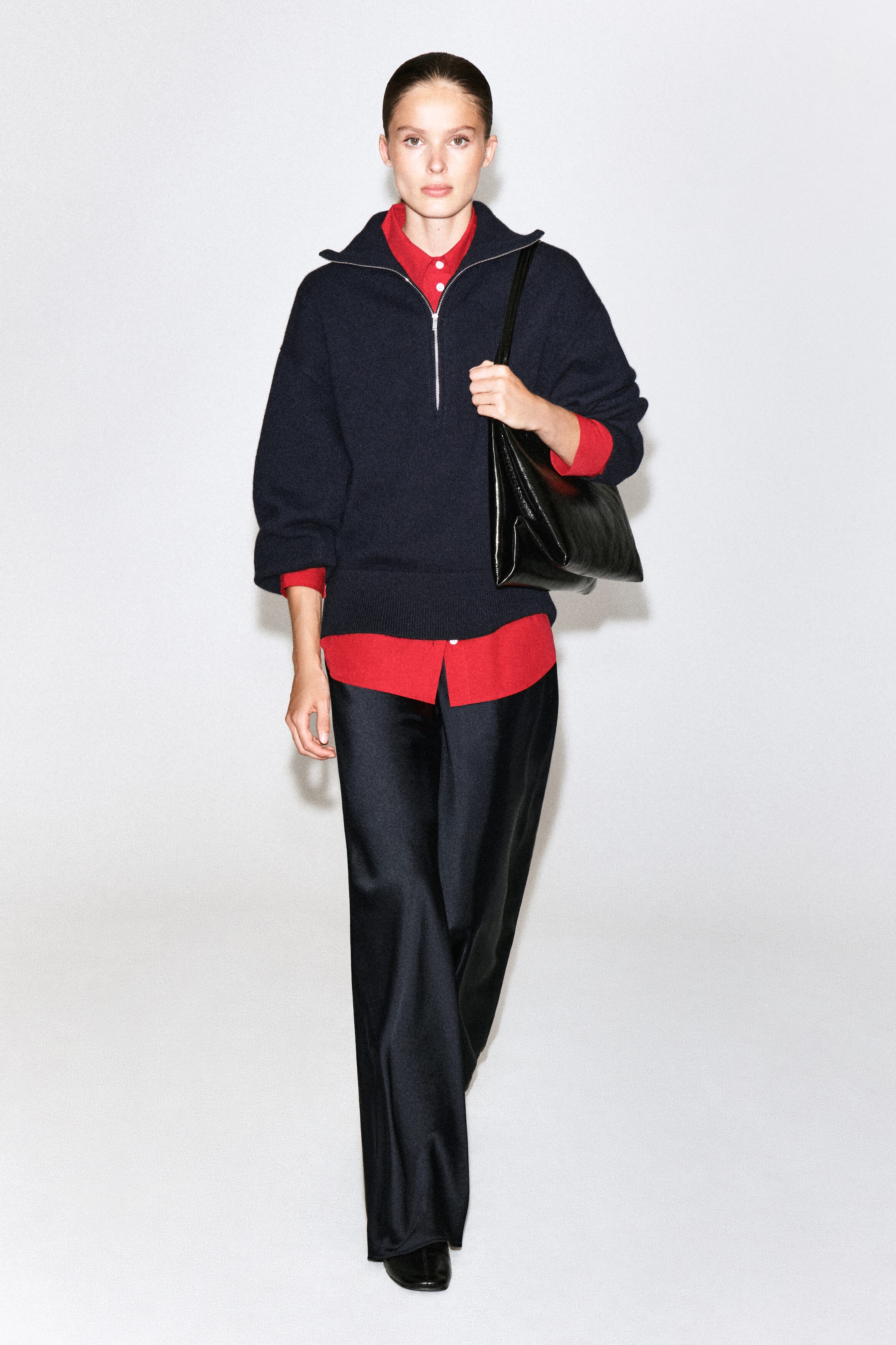 The model wears a navy blue half-zip knitted jumper over a red collared shirt, styled with black wide-leg trousers. The jumper features a high collar with a silver zip, and a black leather shoulder bag is worn over the model's shoulder.