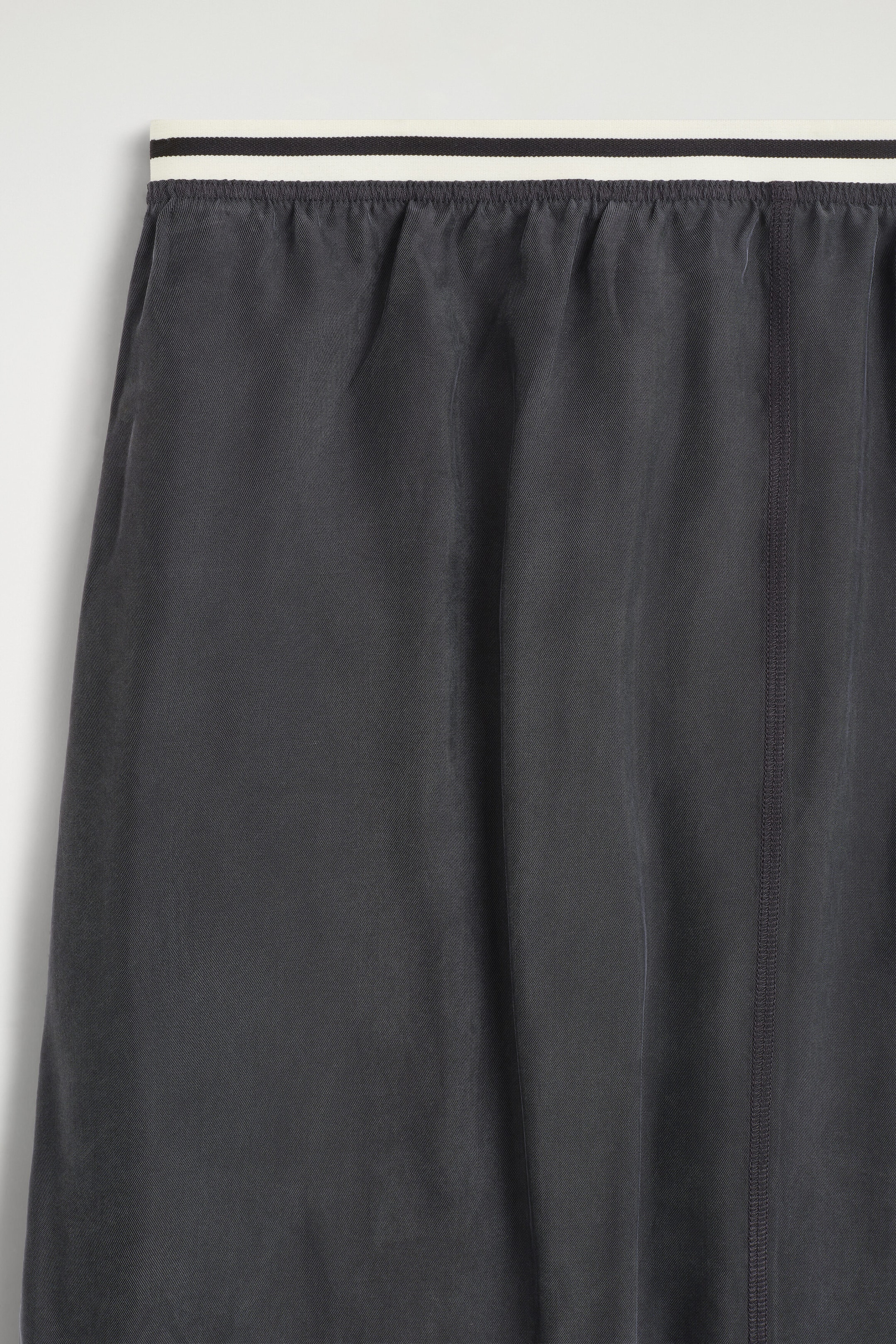 The dark gray garment features a white elastic waistband with thin black stripes. The fabric is gathered beneath the waistband and displays a vertical double-stitched seam on the right side. The dark gray garment features a white elastic waistband with thin black stripes. The fabric is gathered beneath the waistband and displays a vertical double-stitched seam on the right side.