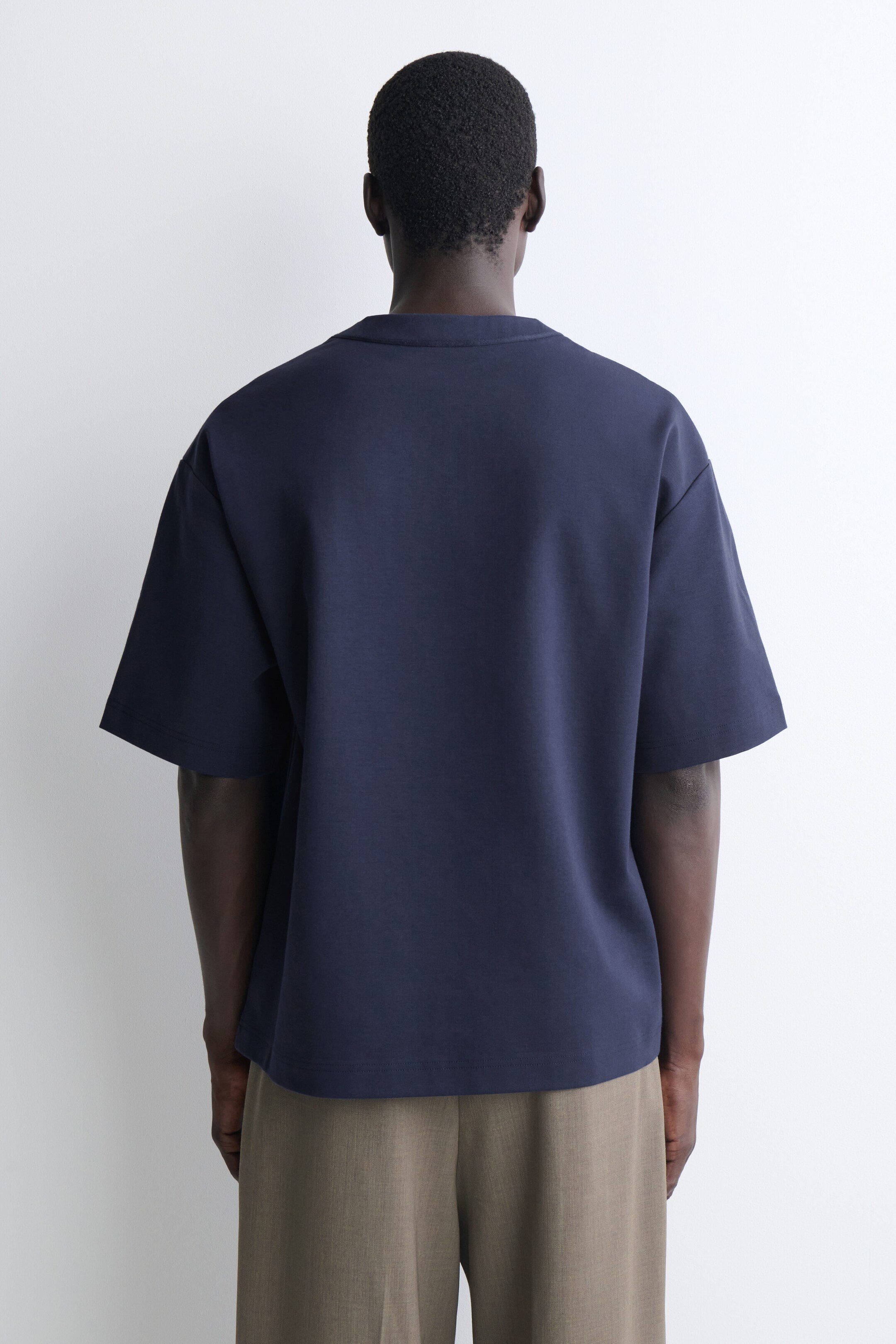 Le t-shirt henley en jersey de coton oversize est présenté de dos, mettant en valeur sa coupe décontractée, ses épaules tombantes et ses manches courtes larges. Le t-shirt bleu marine tombe souplement sur la partie supérieure d’un pantalon vert olive, soulignant son ourlet droit.