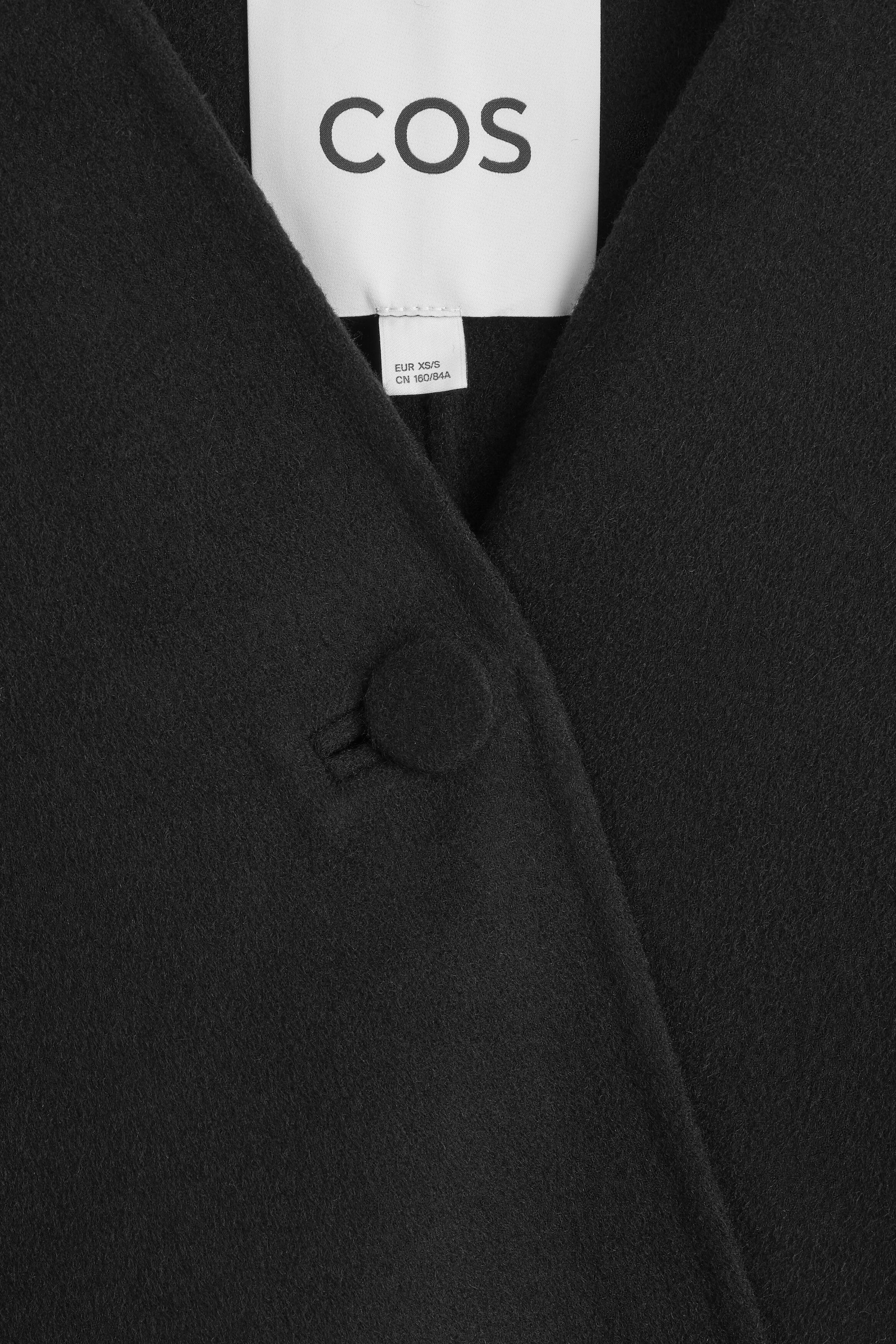 The double-faced wool-blend cape features a close-up of its front neckline, showing a single fabric-covered button closure. The textured black wool-blend fabric displays a white brand label and a smaller size tag at the collar. The double-faced wool-blend cape features a close-up of its front neckline, showing a single fabric-covered button closure. The textured black wool-blend fabric displays a white brand label and a smaller size tag at the collar.