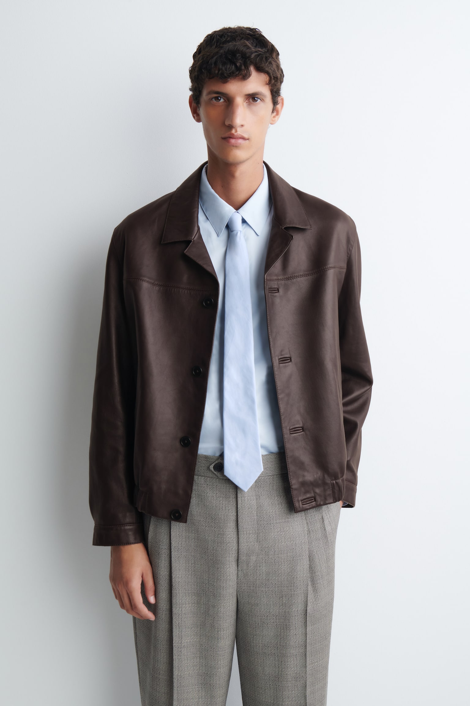 La modelo lleva una corbata de algodón azul claro, anudada bajo una camisa azul claro a juego con cuello. La corbata se combina con una chaqueta de cuero marrón abierta y pantalones plisados grises. - 1