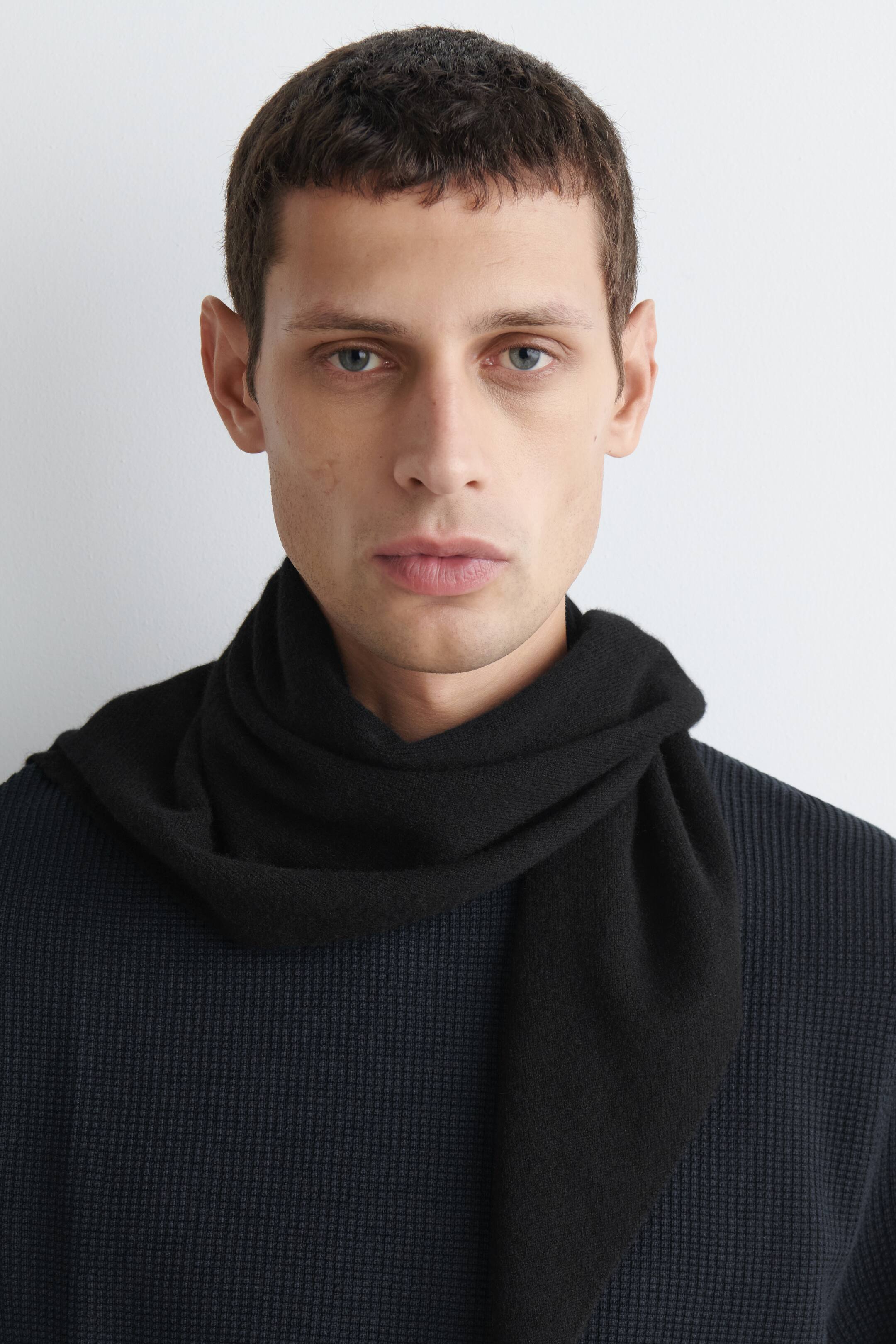 View larger image: A black cashmere triangular scarf is draping loosely around the neck in a front-facing view, with one pointed end hanging down the chest over a dark, textured waffle-knit top.